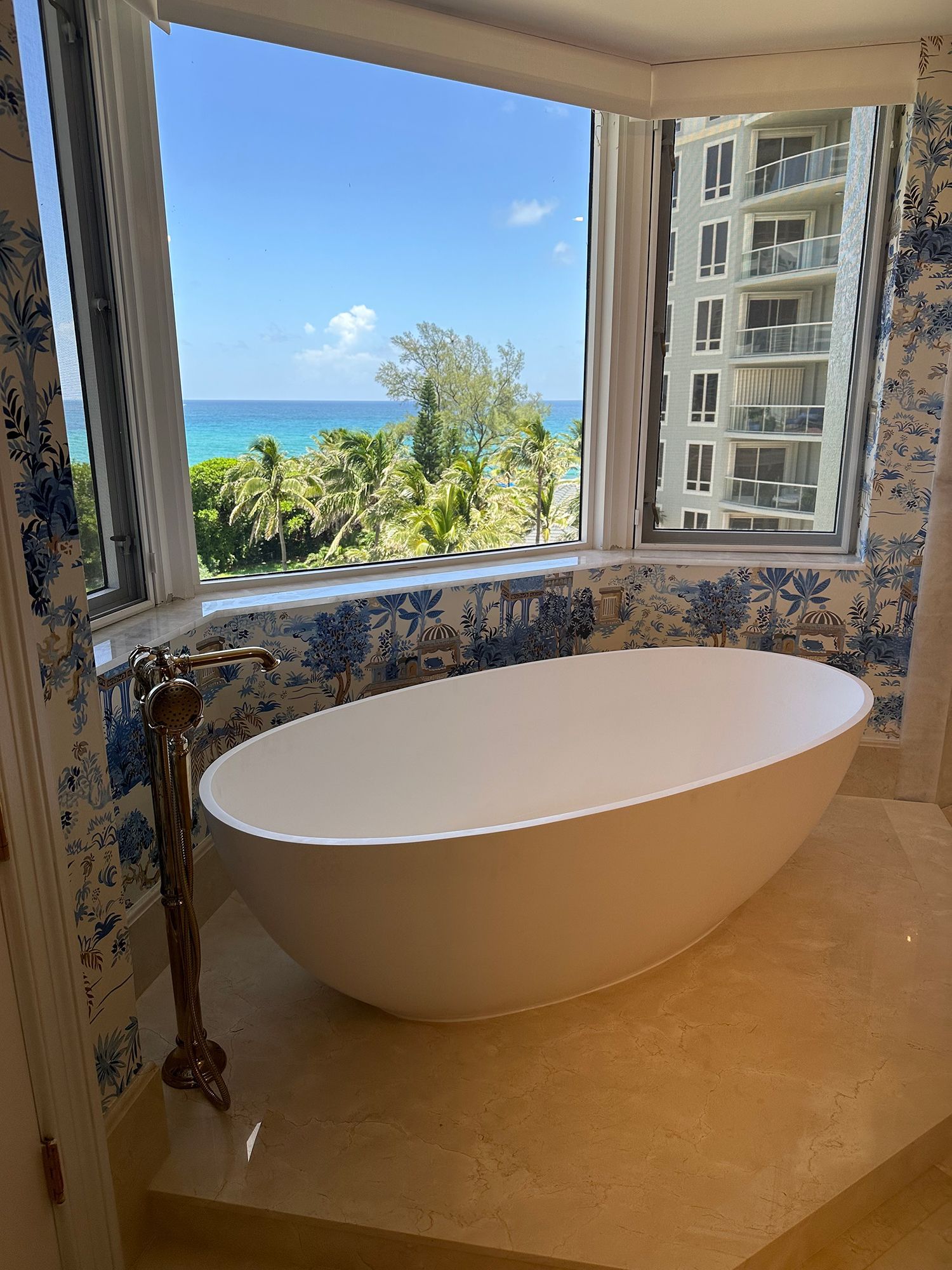 Freestanding white bathtub by window overlooking ocean and palm trees, blue patterned wallpaper.