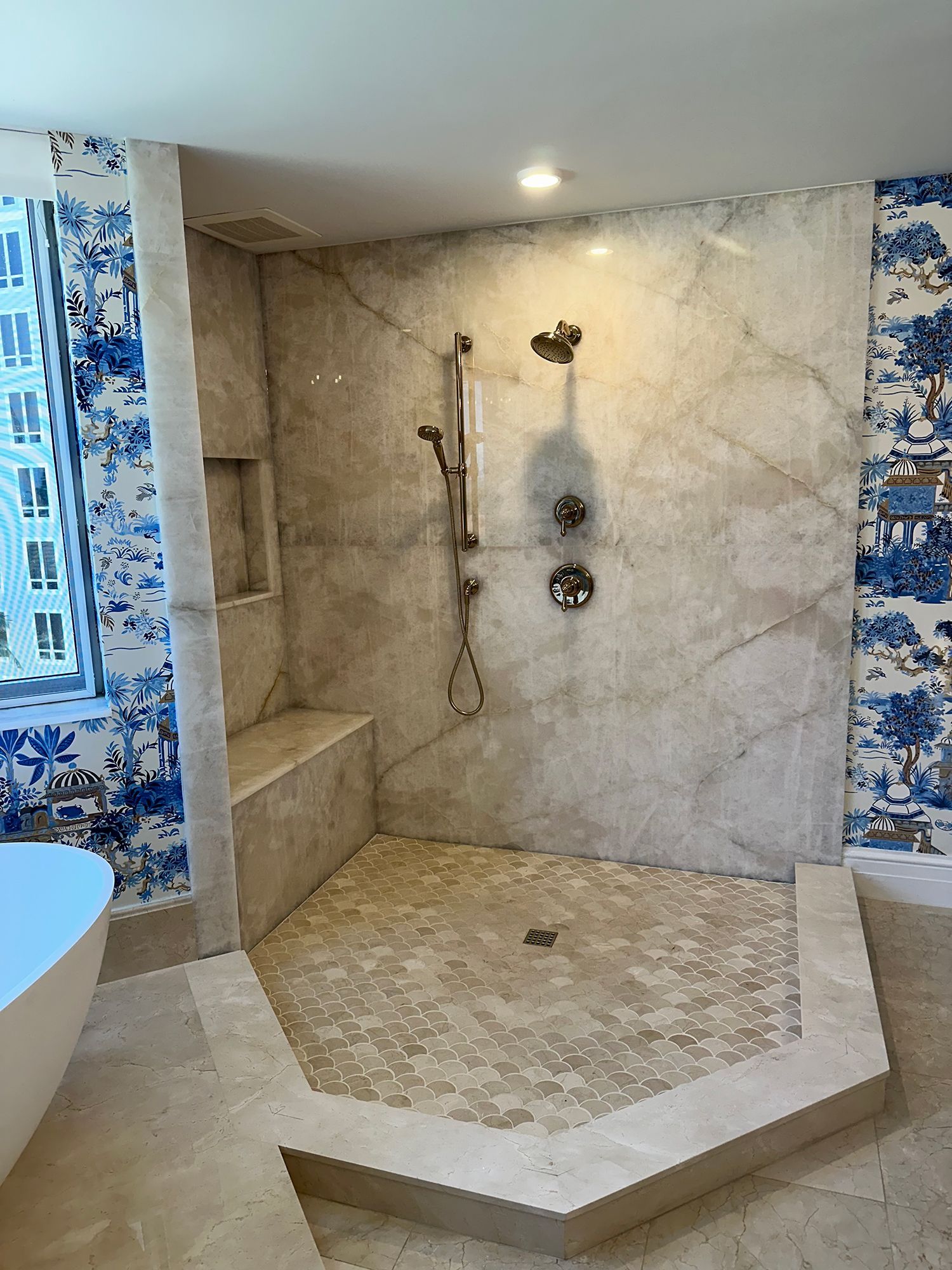 Shower with marble walls, bench, and herringbone floor, with blue and white wallpaper.