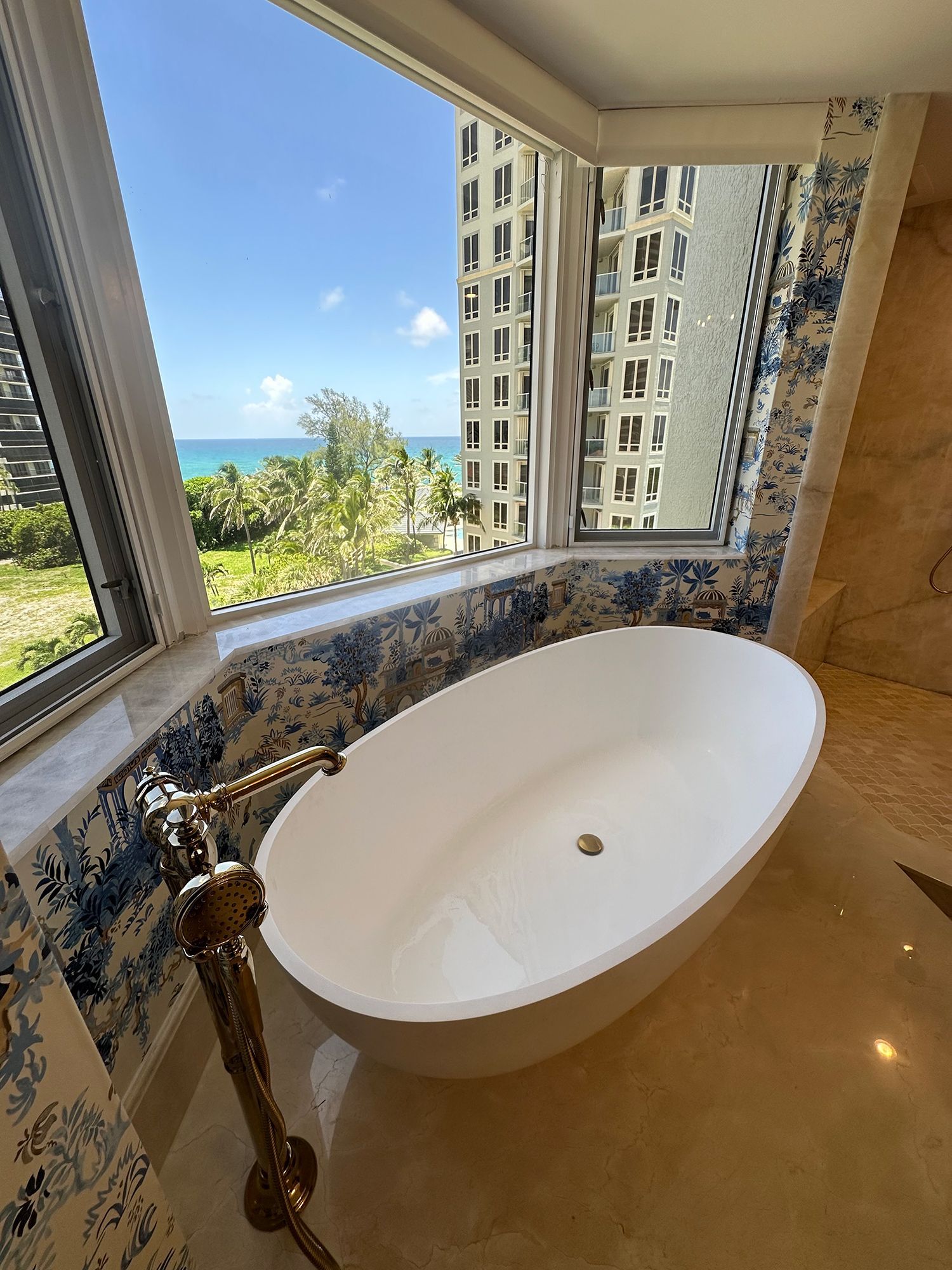 Bathtub by a large window with an ocean view and a tall building in the background; blue and white patterned wall.