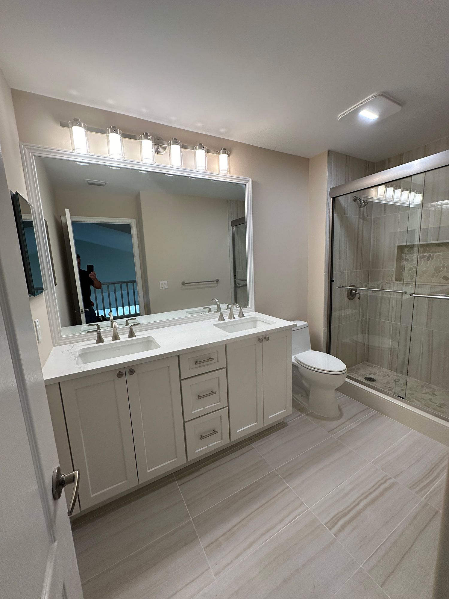 White bathroom with double vanity, large mirror, glass shower, and tile floor.