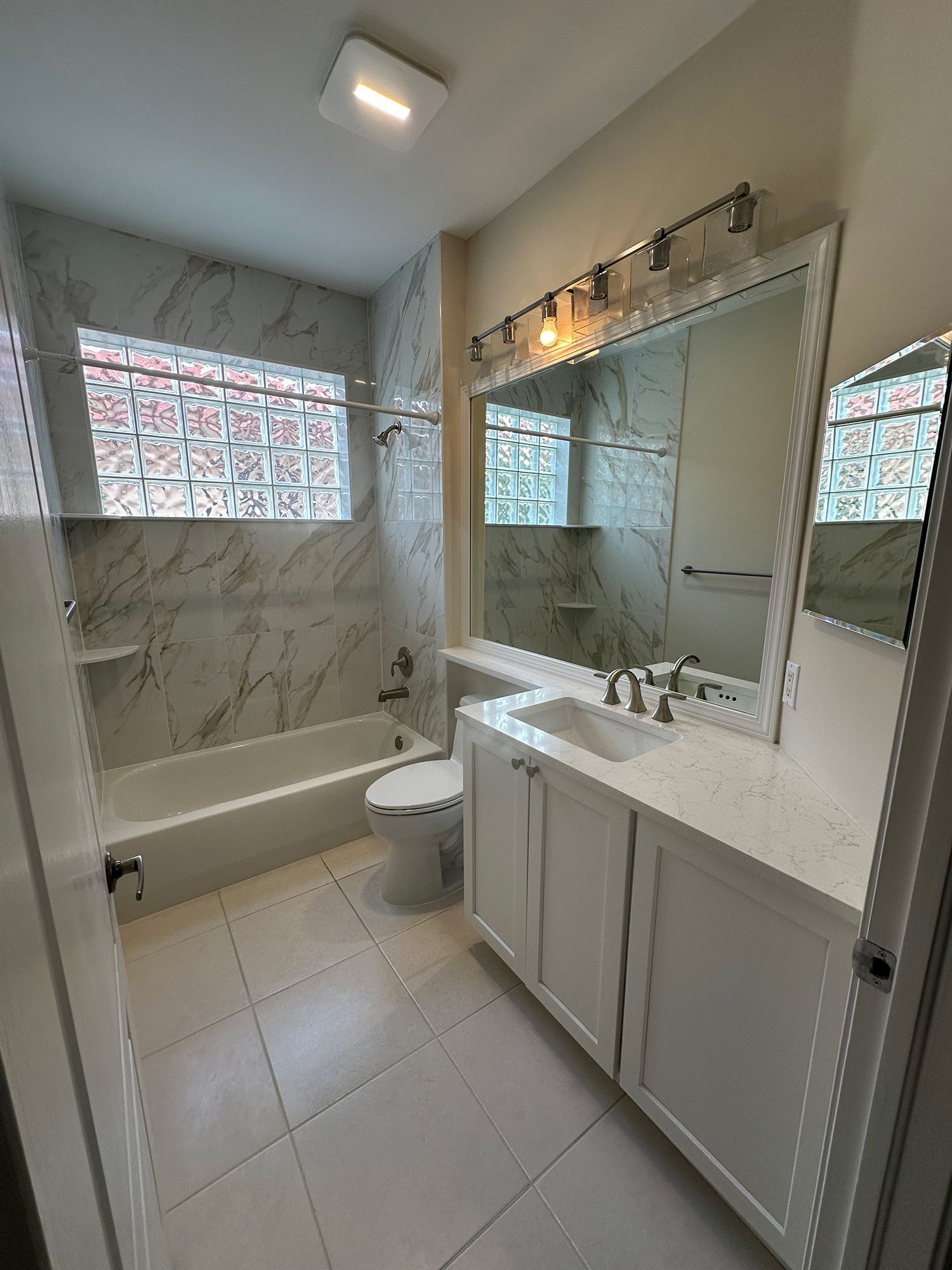 White bathroom with marble-look tile, glass block window, white vanity, tub, and toilet.