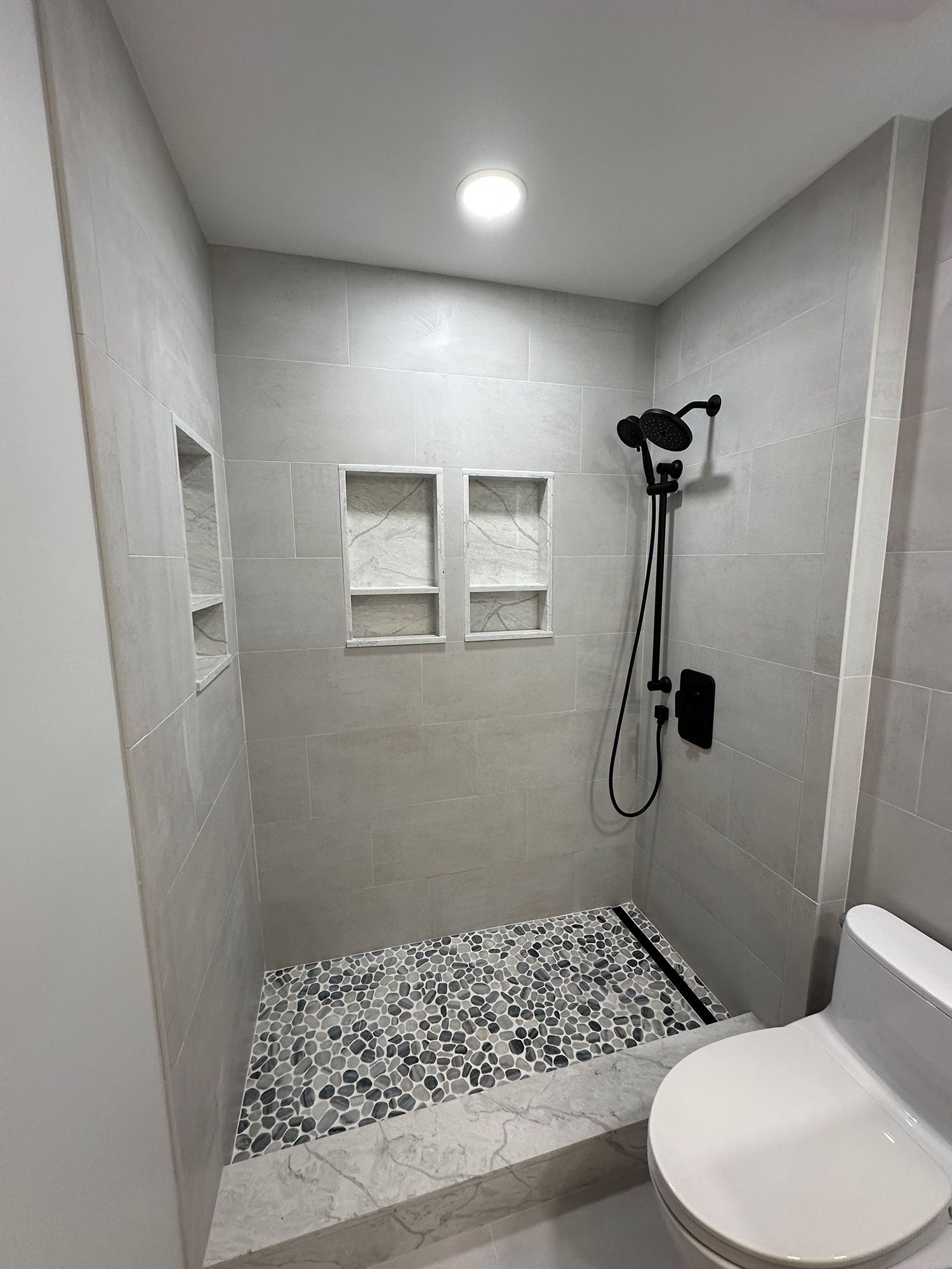 Tiled shower with pebble floor, black shower fixtures, and built-in niches. Toilet visible on the right.