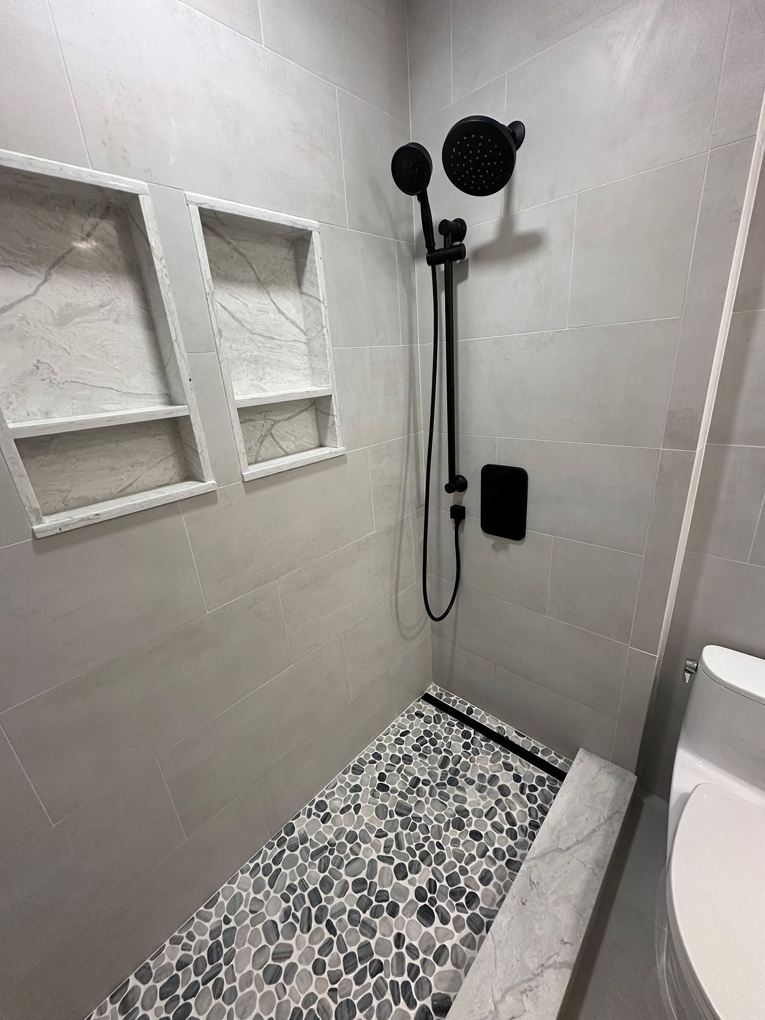 Bathroom shower with gray tiled walls, pebble floor, black fixtures, and built-in shelves.