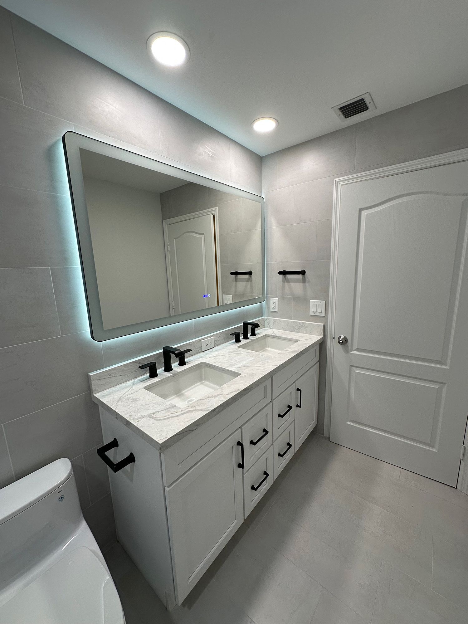 Modern bathroom with white vanity, marble countertop, black fixtures, lit mirror, and gray tiled walls.