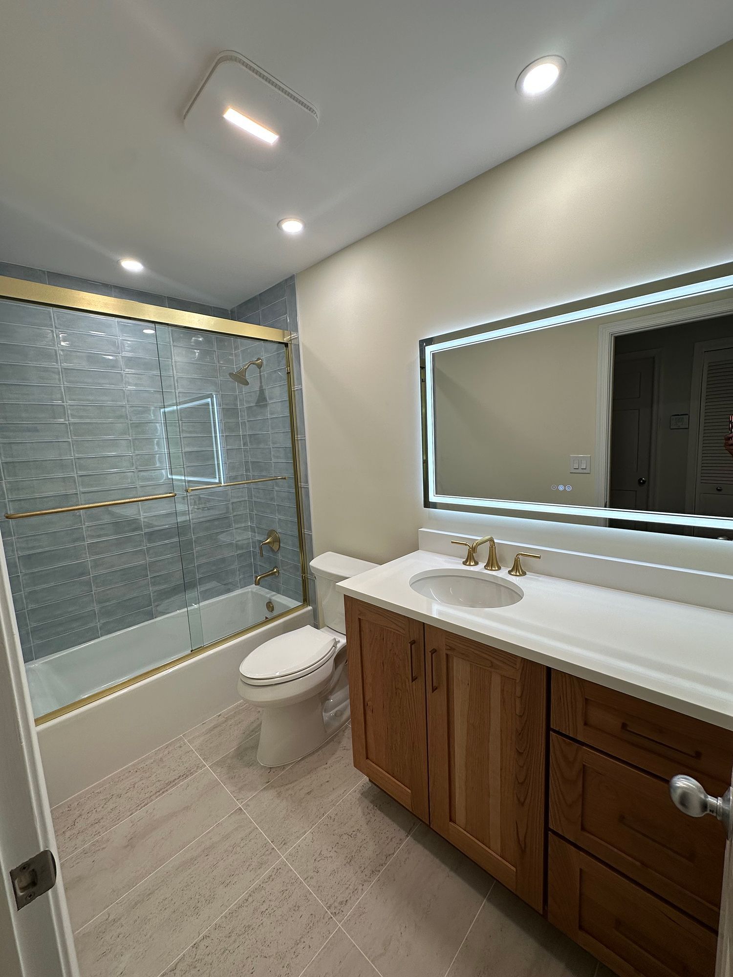 Bathroom with a blue tiled shower, wood vanity, and a lit mirror.