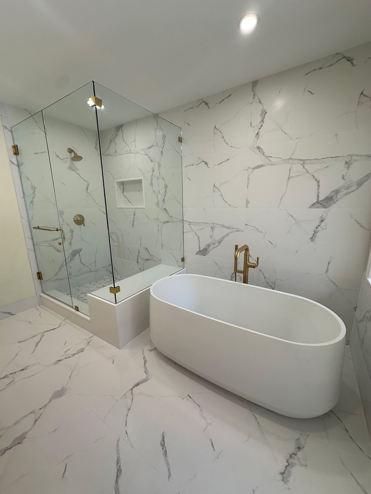 Modern bathroom with marble-look tile, glass shower, freestanding white tub, and gold fixtures.