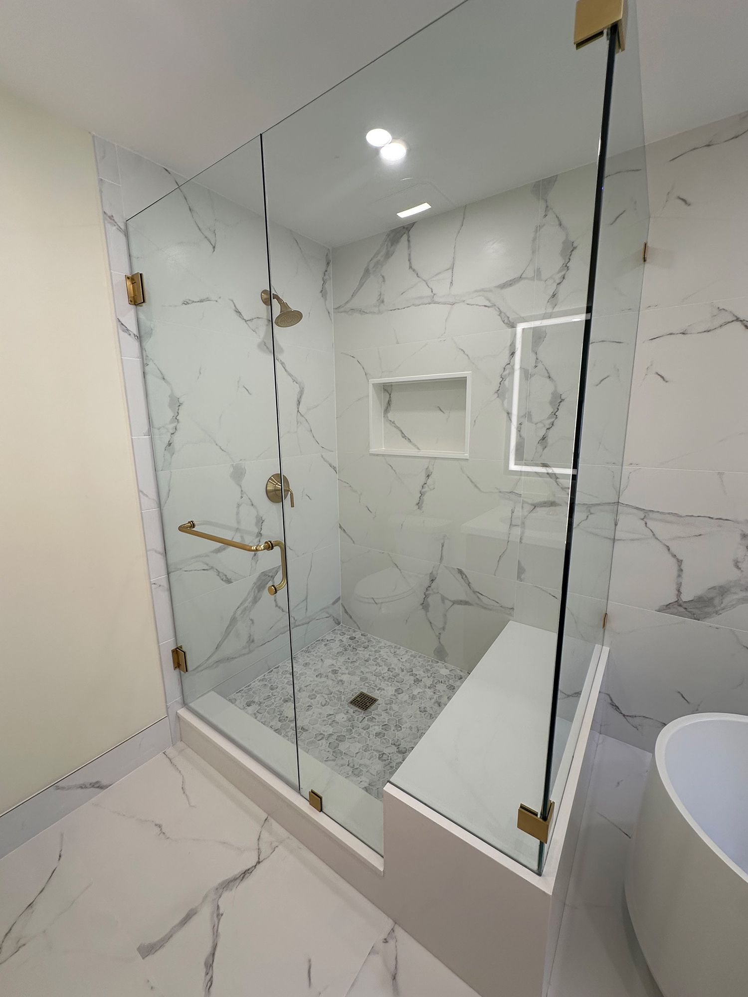 Modern marble-tiled shower with glass doors, gold fixtures, built-in bench, and pebble floor.