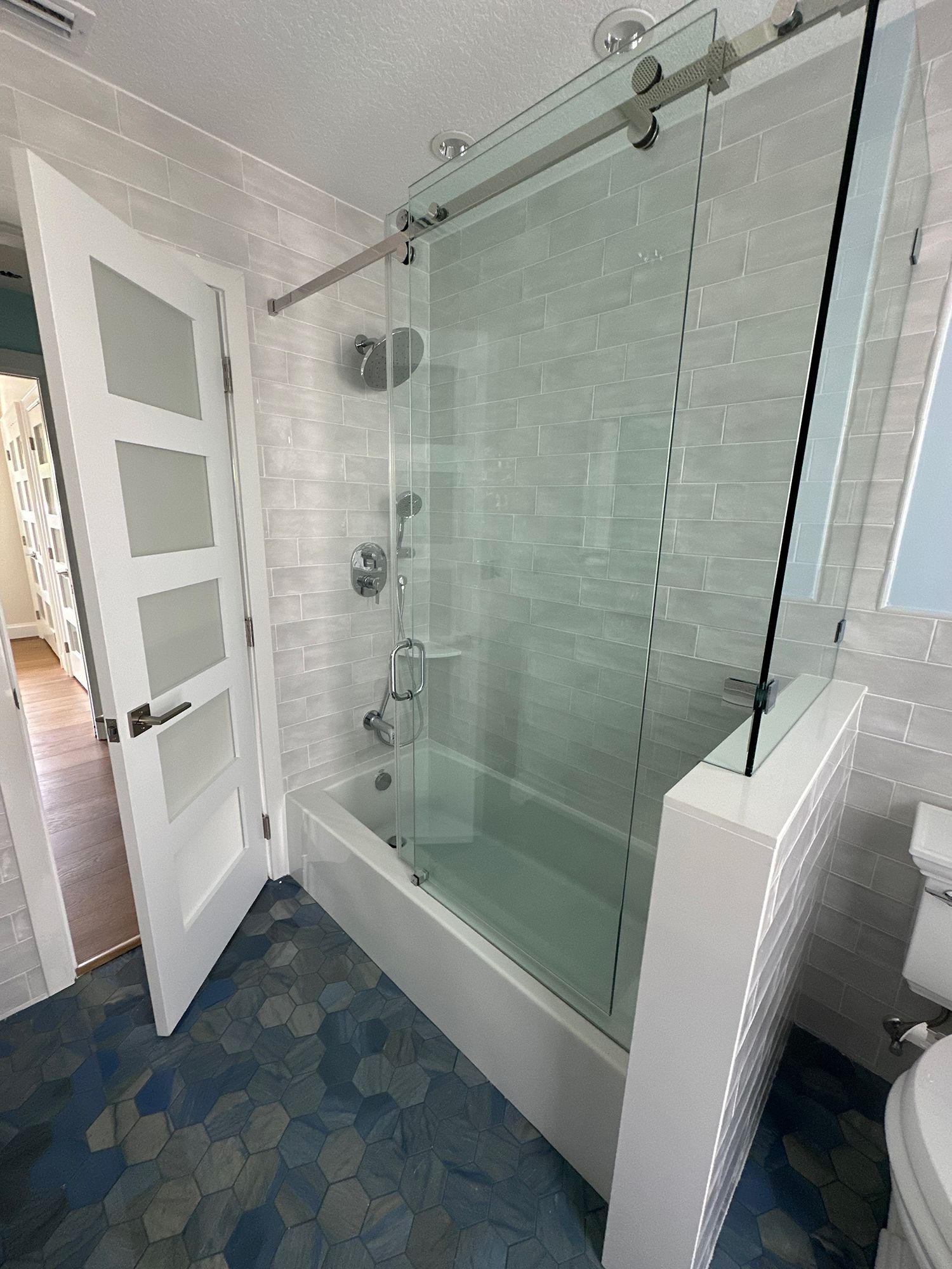 Bathroom with glass shower door, white tub, blue tile floor. Open white door to hallway.