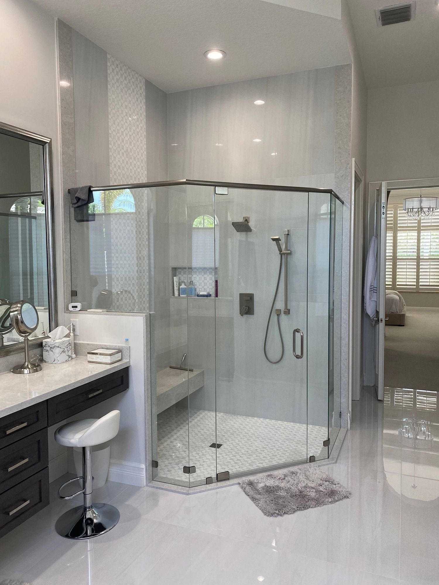 Modern bathroom with a glass-enclosed shower, vanity with stool, and white tile flooring.