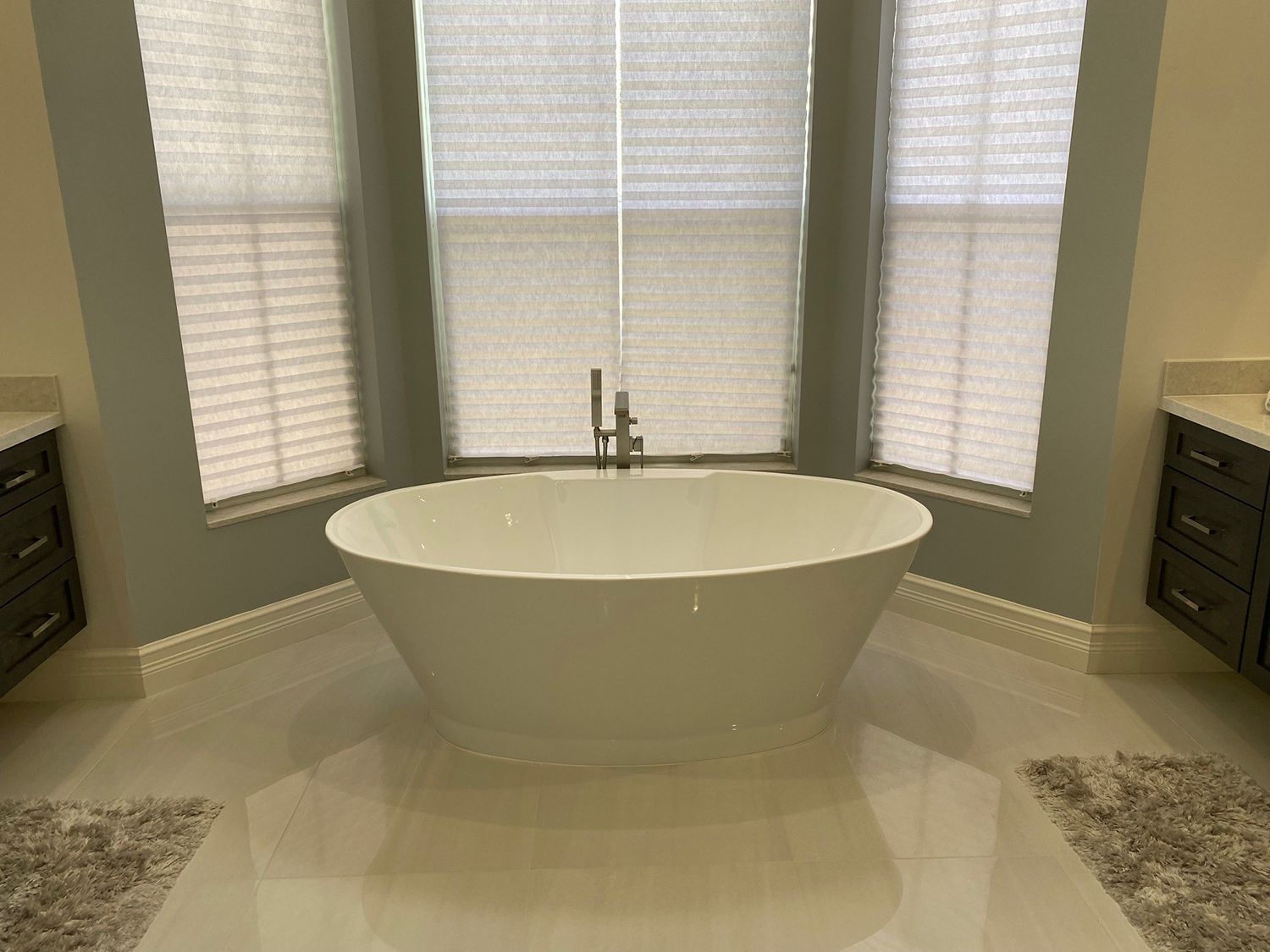 White bathtub in front of a window with blinds, flanked by dark cabinets in a bathroom.