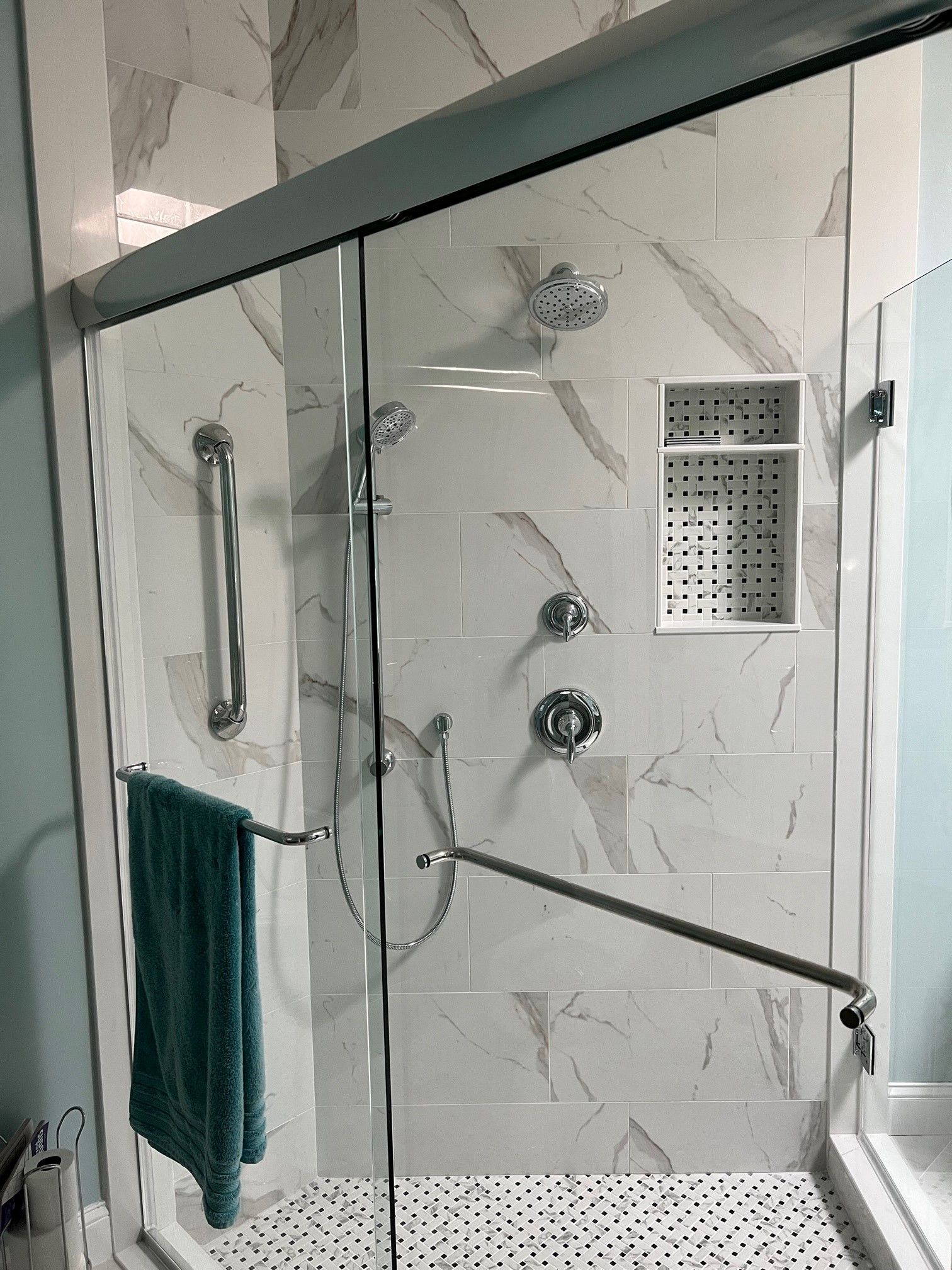 Modern shower with marble-look tile, glass doors, and grab bars. Teal towel hangs on a bar.