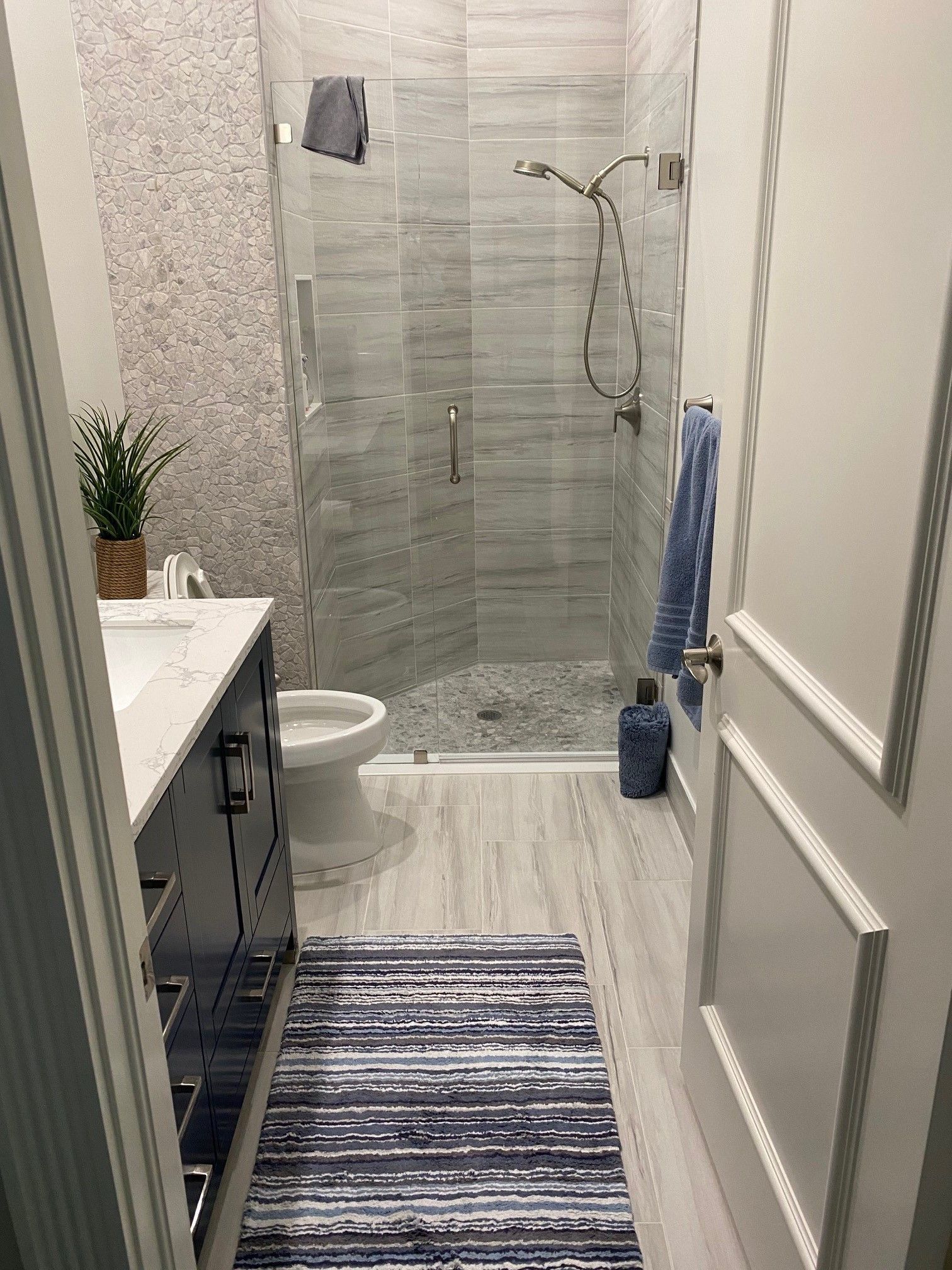 Bathroom with gray tiled shower, vanity, and blue striped rug. Light wood-look flooring.