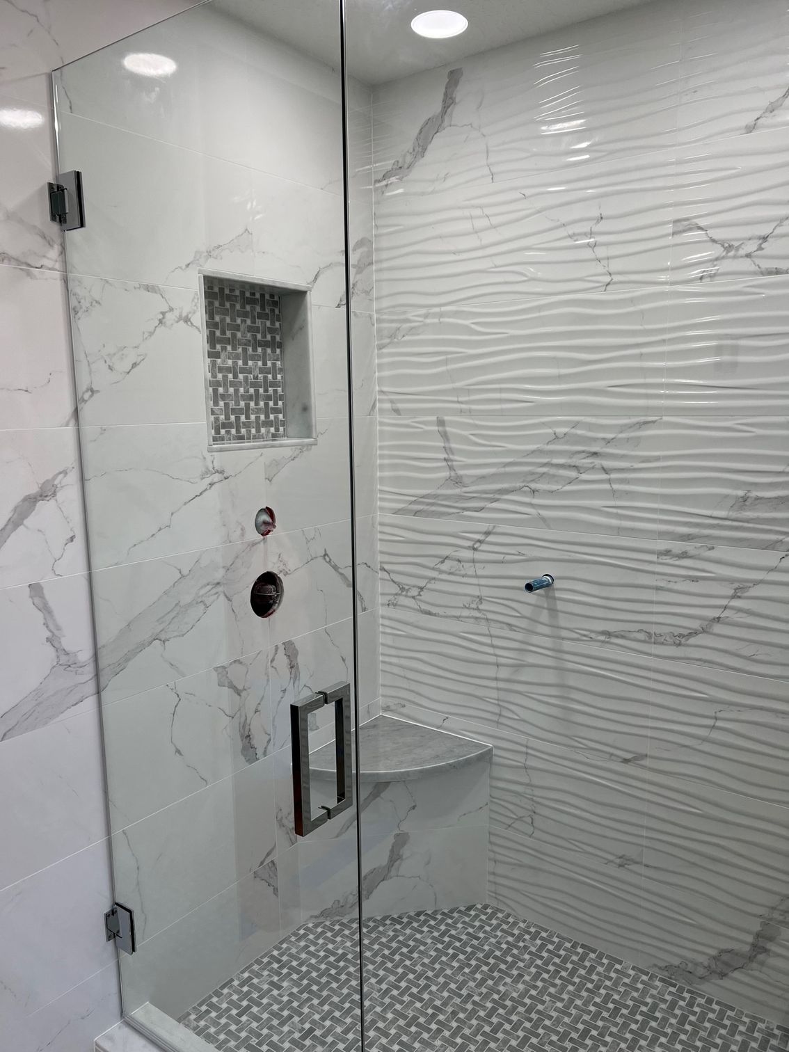 Modern shower with glass door, white marble and wavy tile, gray mosaic floor, and built-in bench.