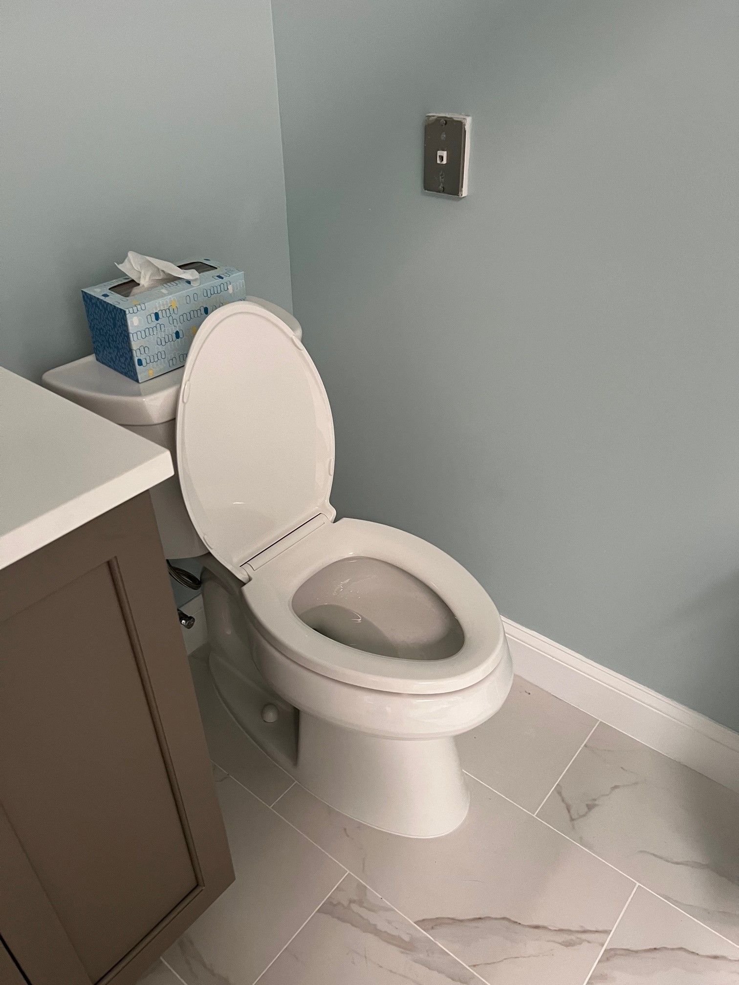 Toilet in a bathroom with blue walls, tissue box, and light-colored tiled floor. Toilet seat is up.