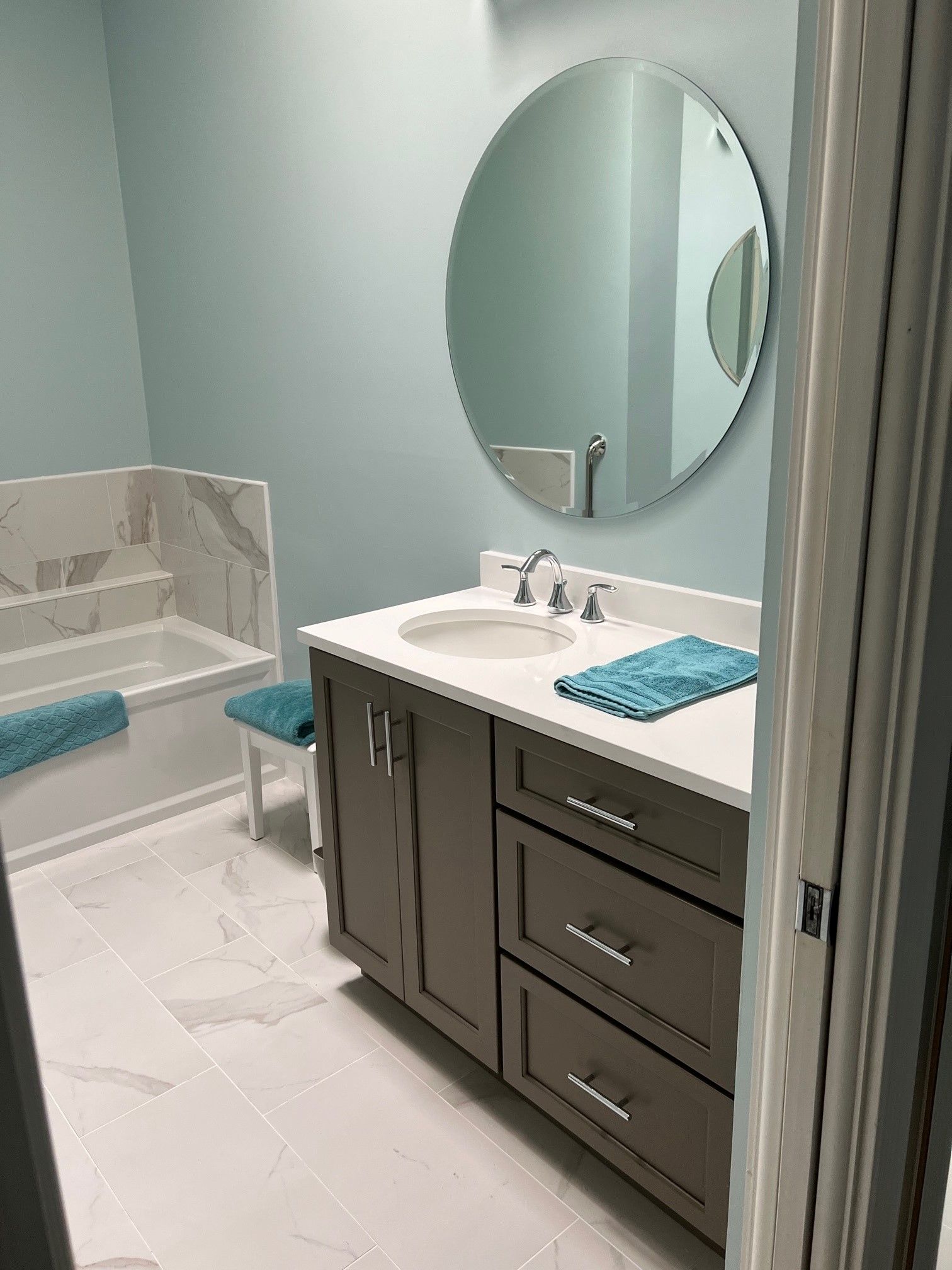 A remodeled bathroom with light blue walls, a gray vanity, and a large round mirror.