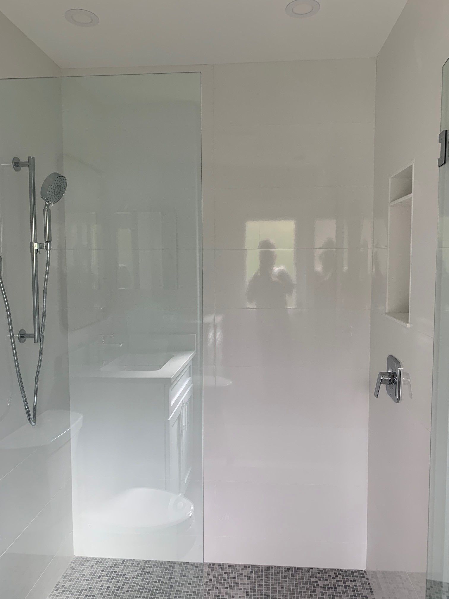 Modern white bathroom with a glass-enclosed shower, showerhead, and a built-in shelf. A person's reflection is visible.