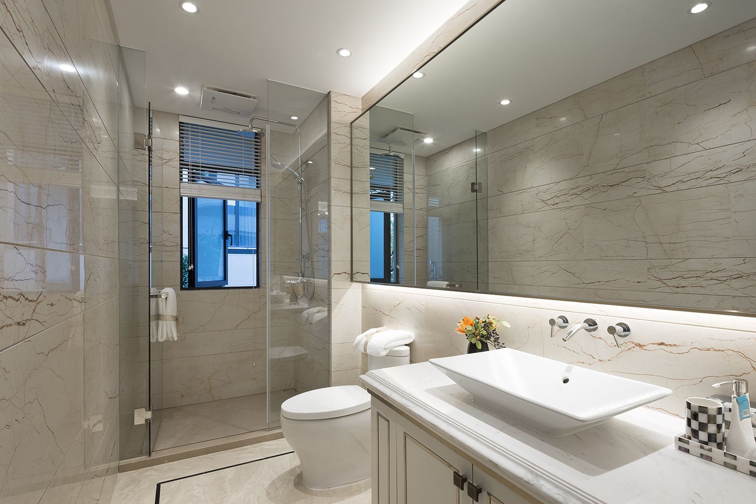 Modern bathroom with marble walls, glass shower, toilet, sink, and large mirror.