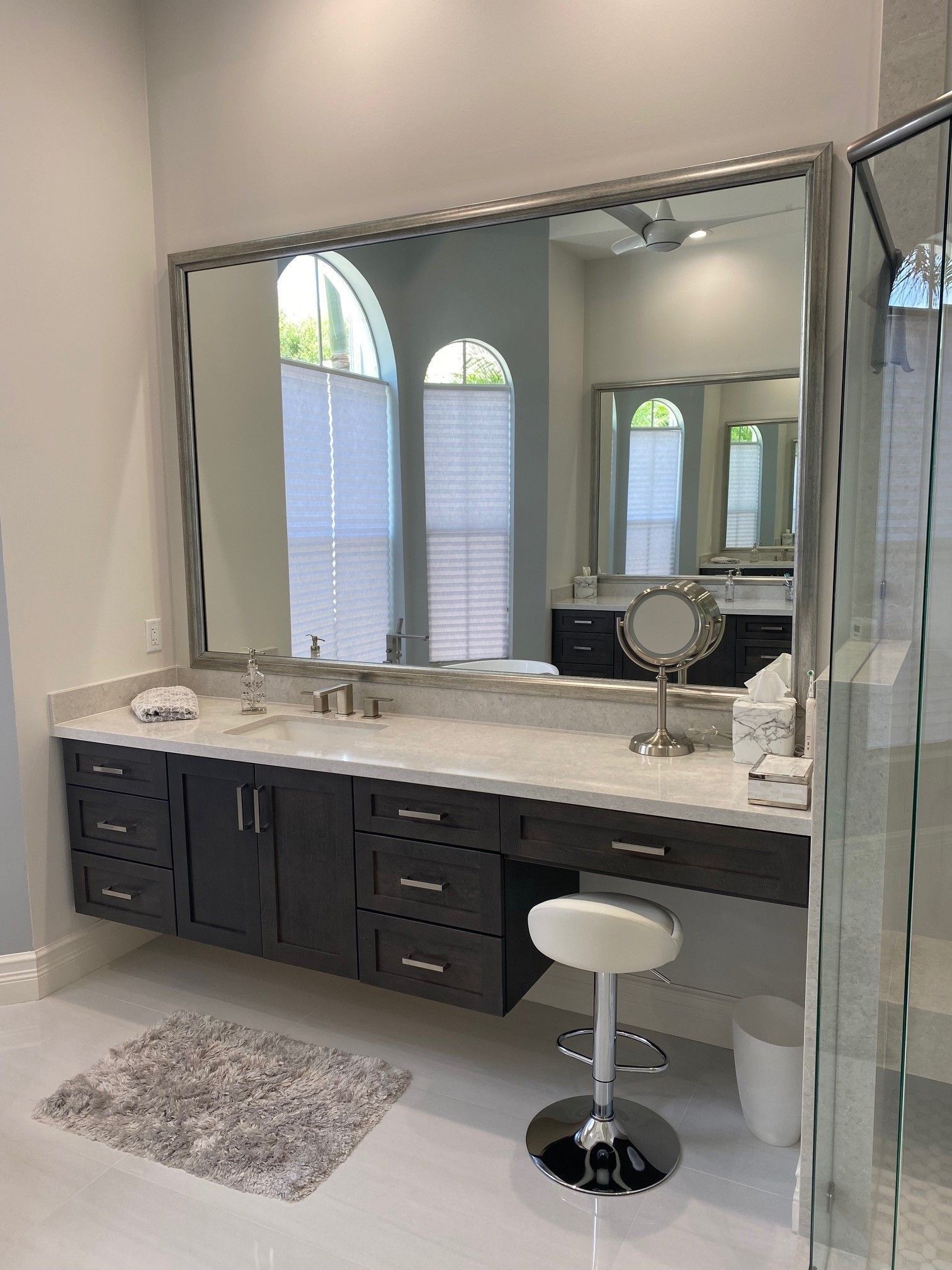 Bathroom vanity with large mirror, dark cabinets, white countertop, and a stool.