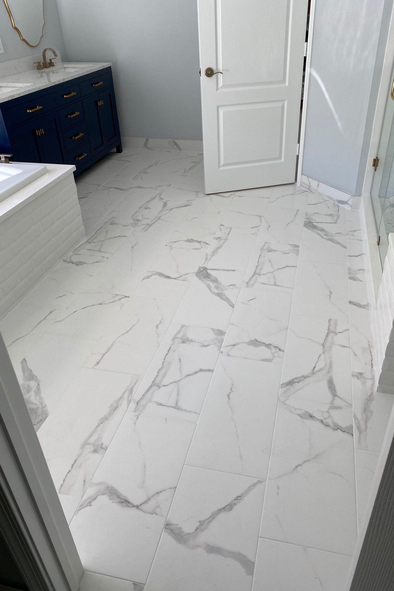 White marble-look bathroom floor with gray veining. Navy vanity, white door, and tiled tub are visible.