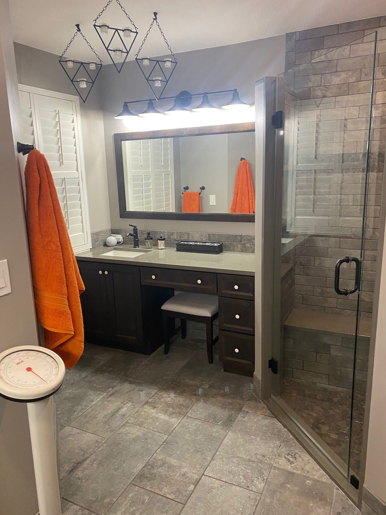 Bathroom with vanity, mirror, shower, and weight scale. Gray and brown tones, orange towel.