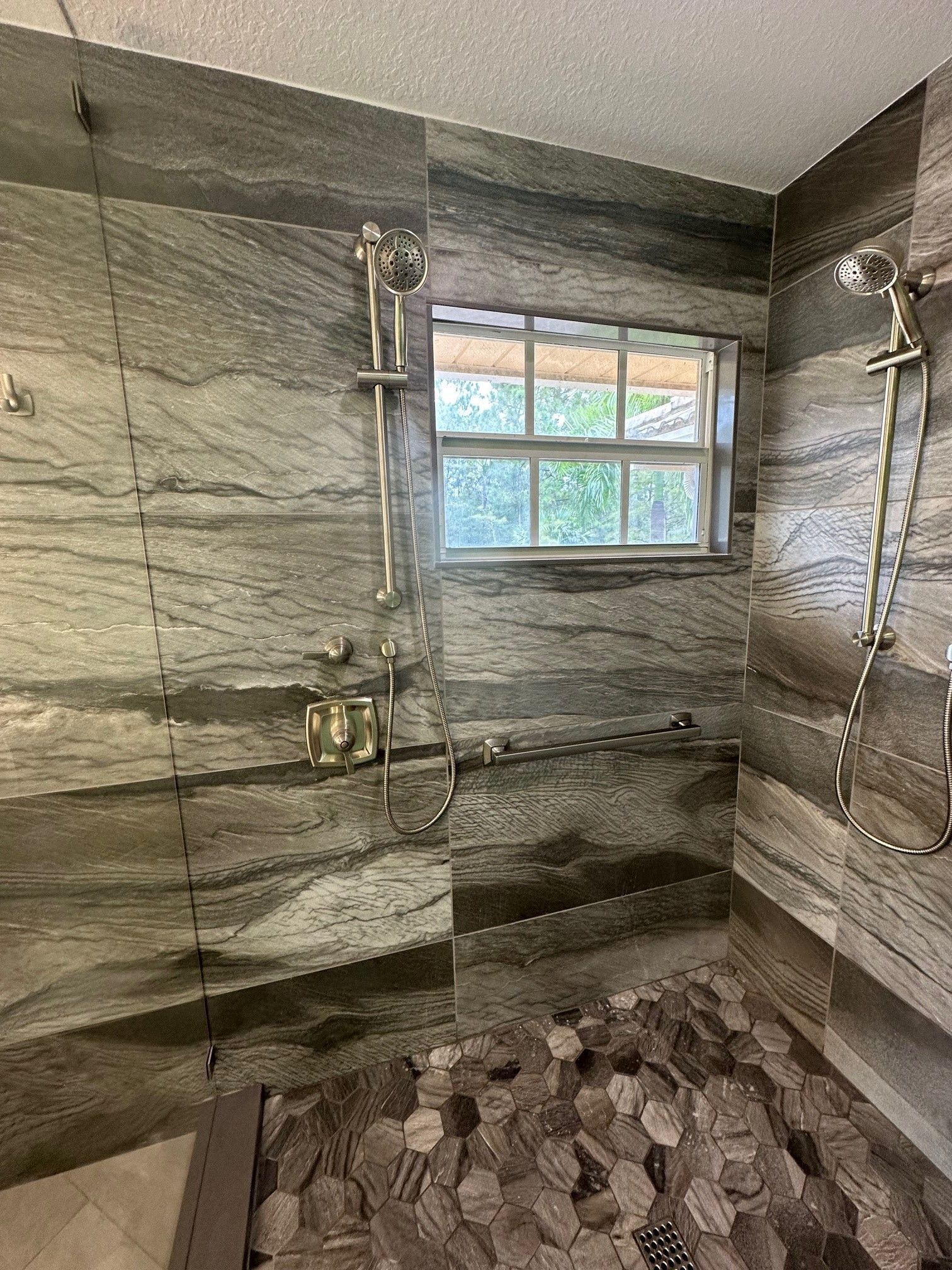 Shower with grey-toned stone tile walls, pebble floor, and a window. Two shower heads and a soap rack are visible.