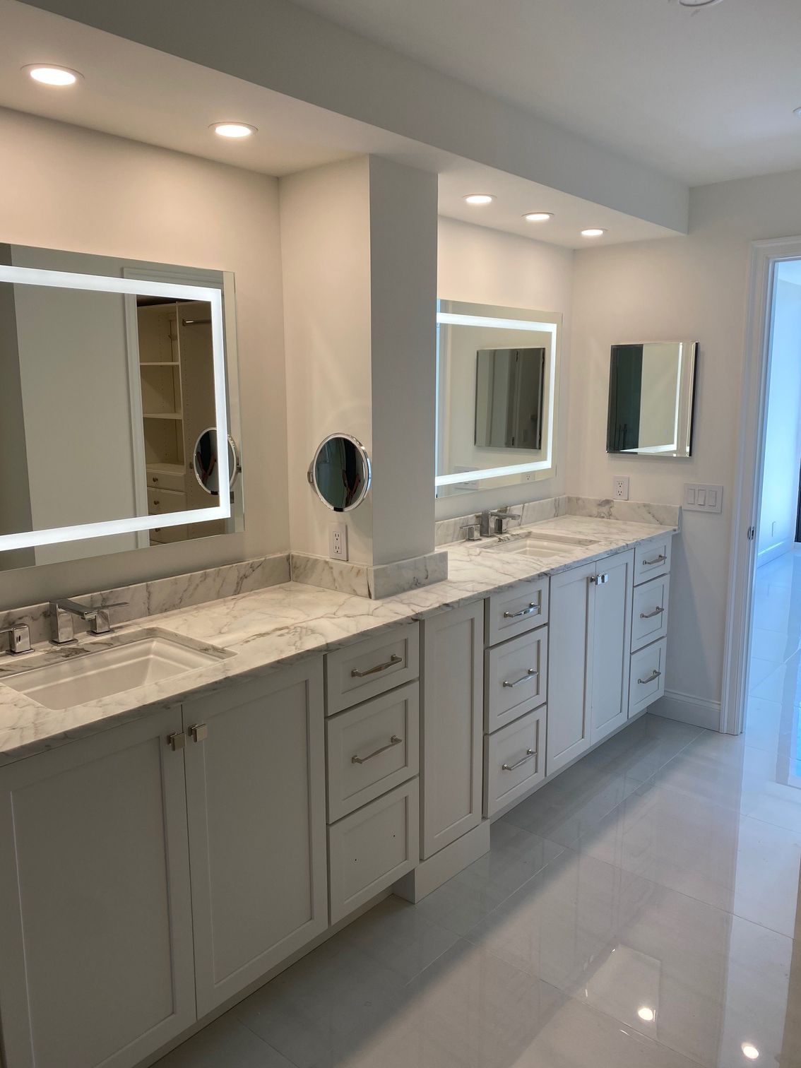 Modern white bathroom with double vanity, lighted mirrors, and white flooring.