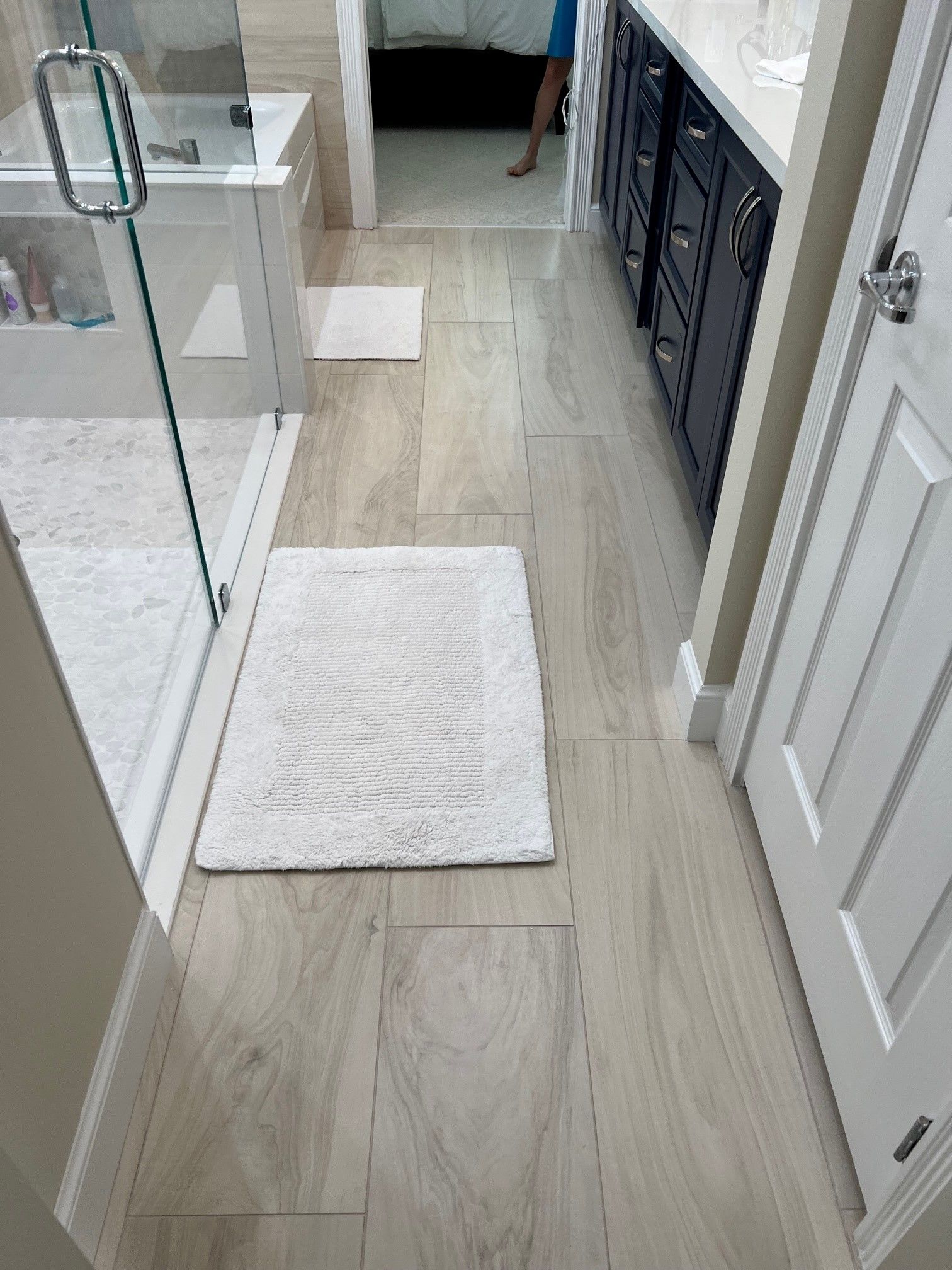 Bathroom with white rug on floor, shower, dark cabinet, white door.