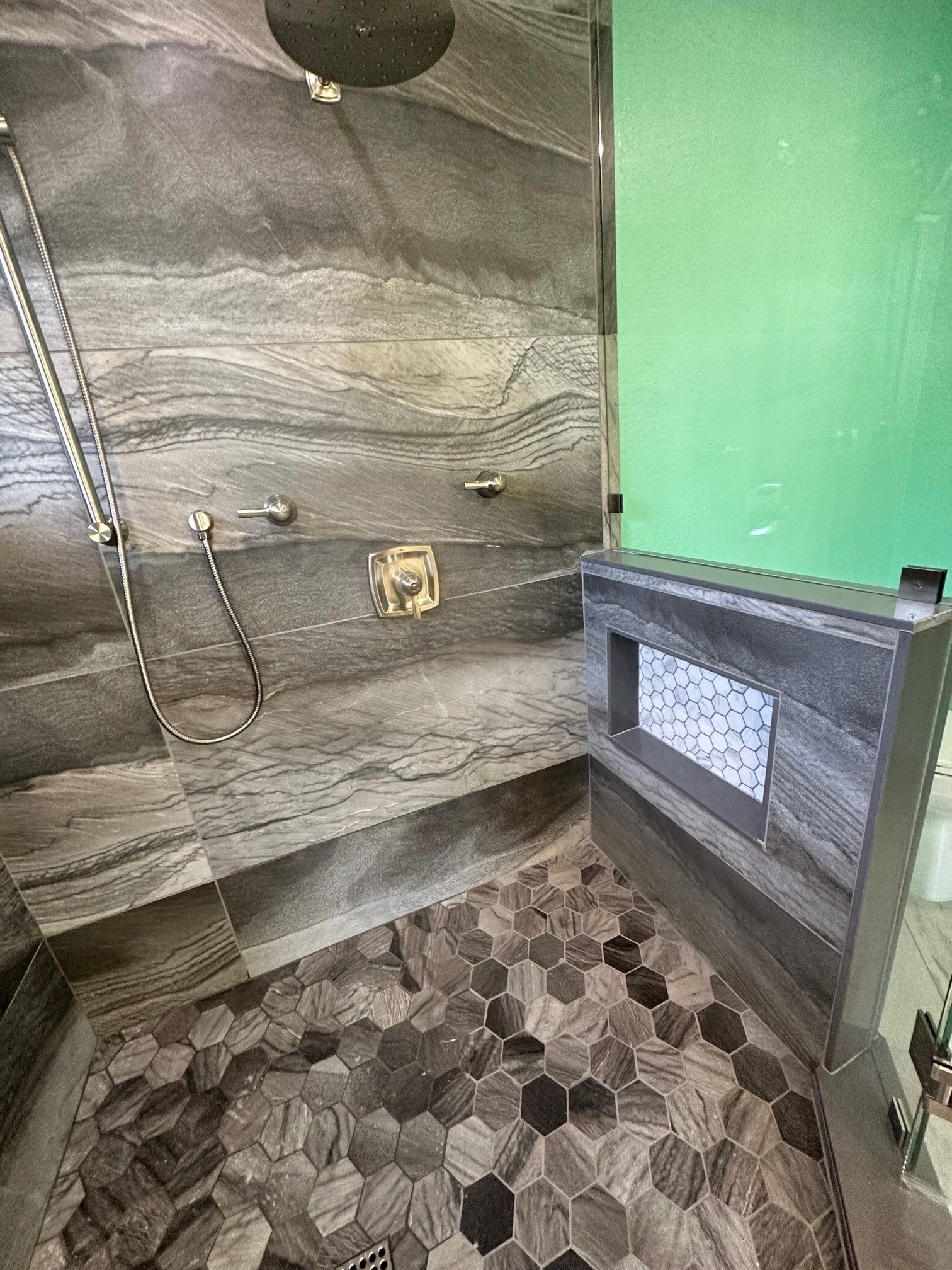 Shower with grey and black stone tile walls and a hexagon tile floor; rain shower head, handheld shower, and a built-in shelf.