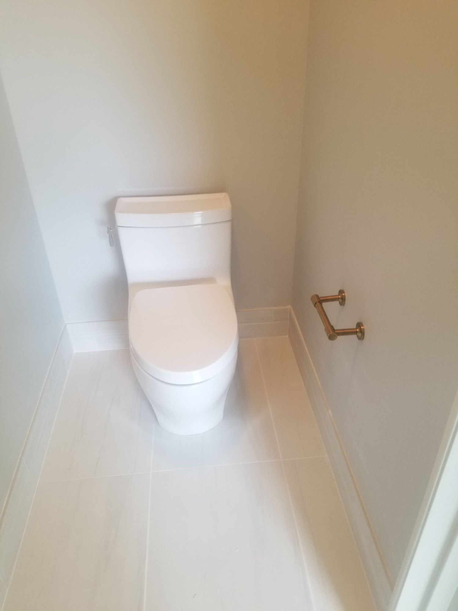 White toilet in a small bathroom with light-colored tile flooring and brass water pipes on the wall.