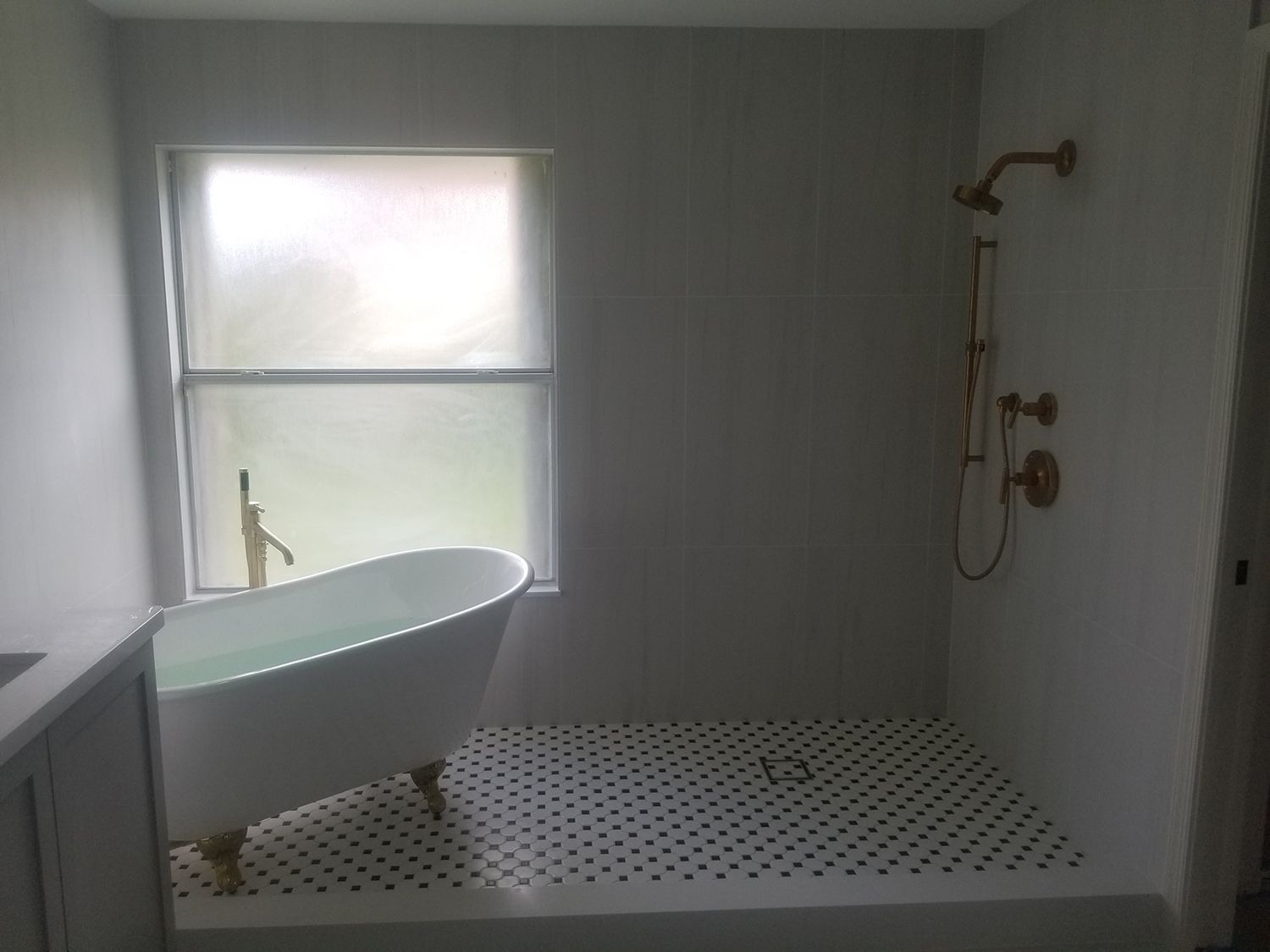 Bathroom with white clawfoot tub under window, gold shower fixtures, and black and white tile floor.