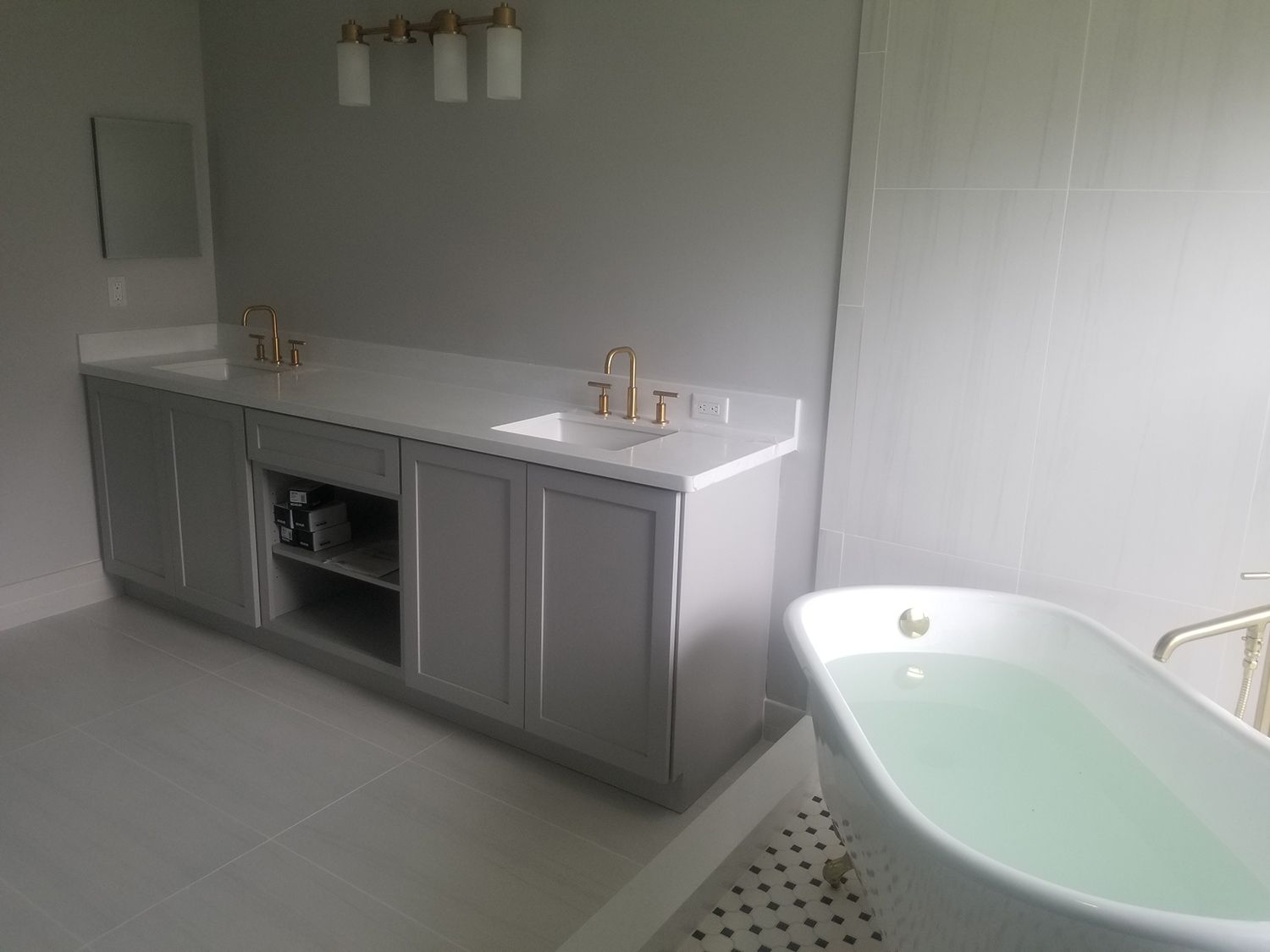 Bathroom with gray vanity, gold fixtures, white countertop, and freestanding tub filled with water.
