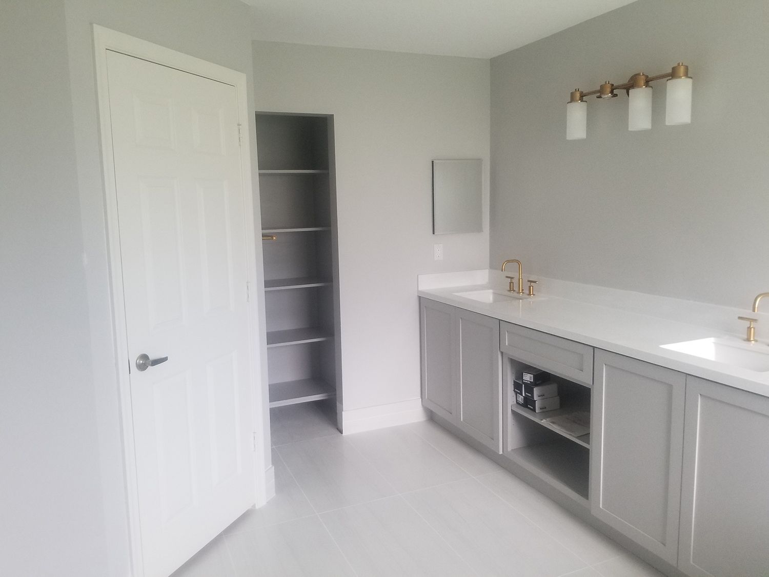 Bathroom with white door, gray cabinets, double sinks, and a storage area.