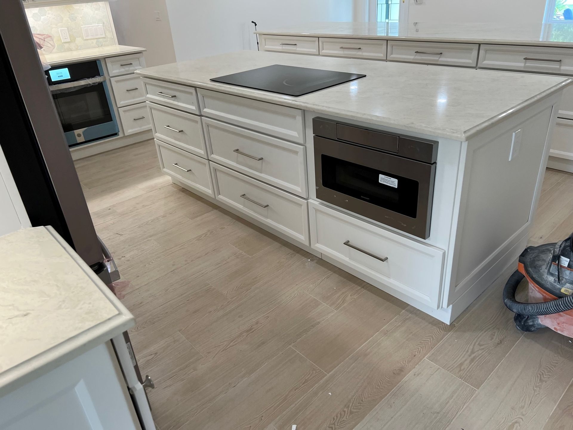 White kitchen island with cooktop and built-in microwave, surrounded by drawers and cabinets, on light wood flooring.