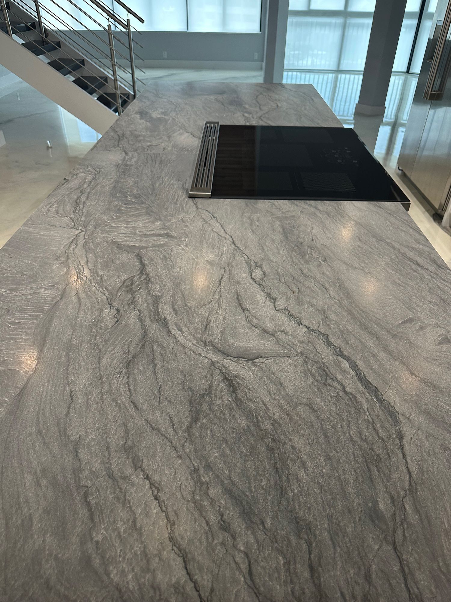 Gray stone kitchen island with cooktop, near stairs.