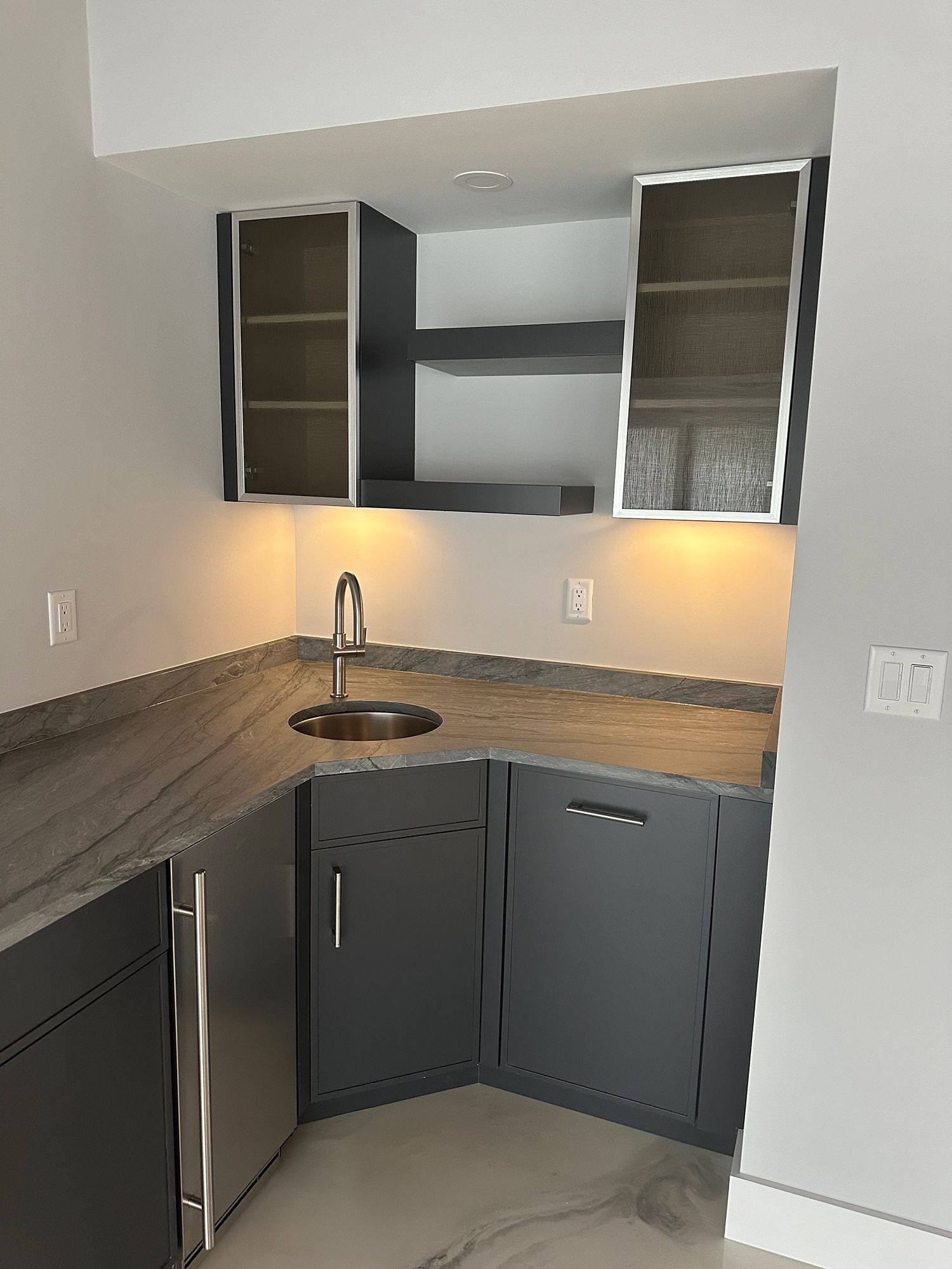 A modern, corner wet bar with gray cabinets, countertop, and sink. Overhead, a shelf and storage.