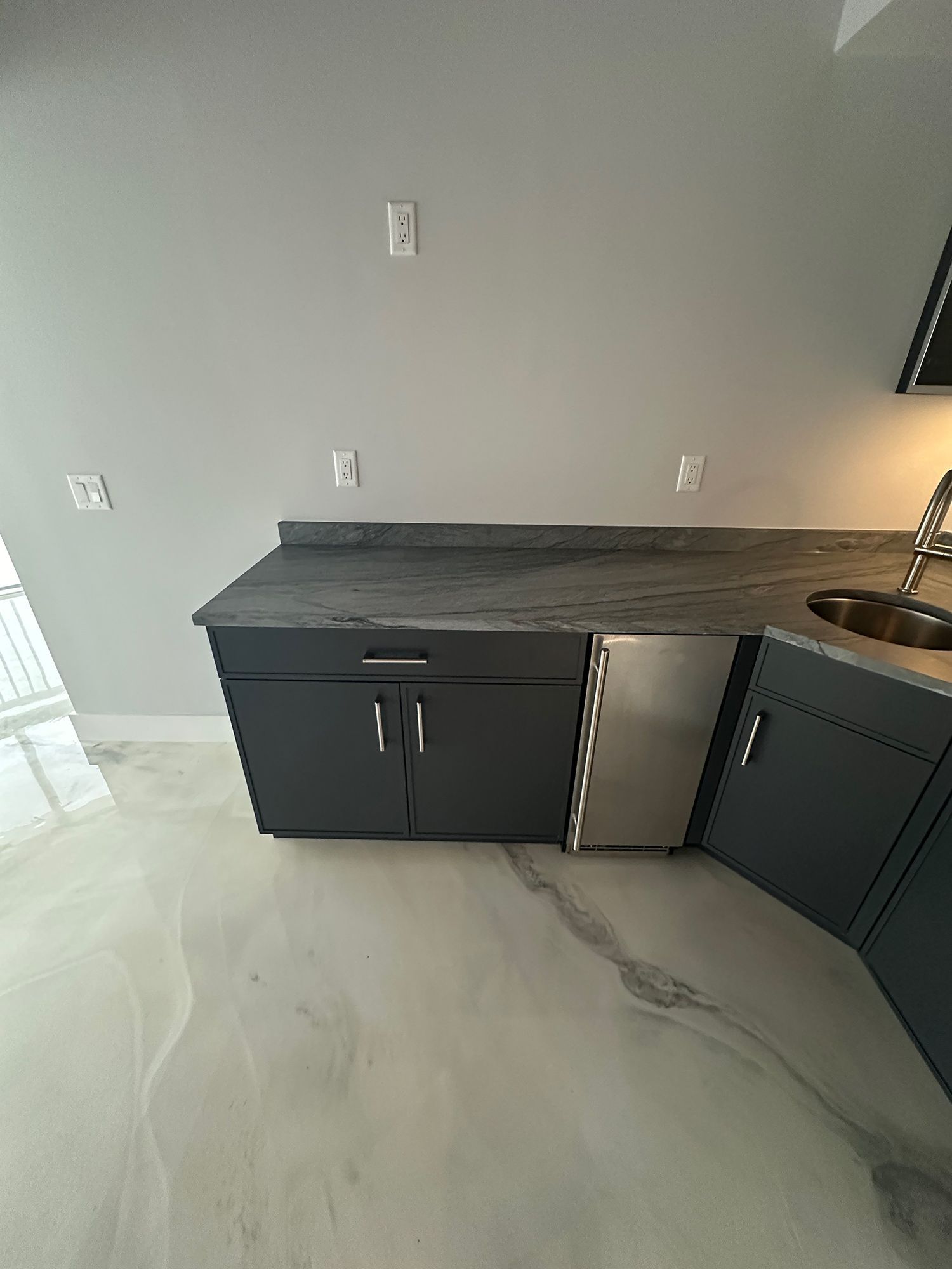 Dark gray cabinets with a countertop, mini-fridge, and sink against a gray wall, on a reflective floor.