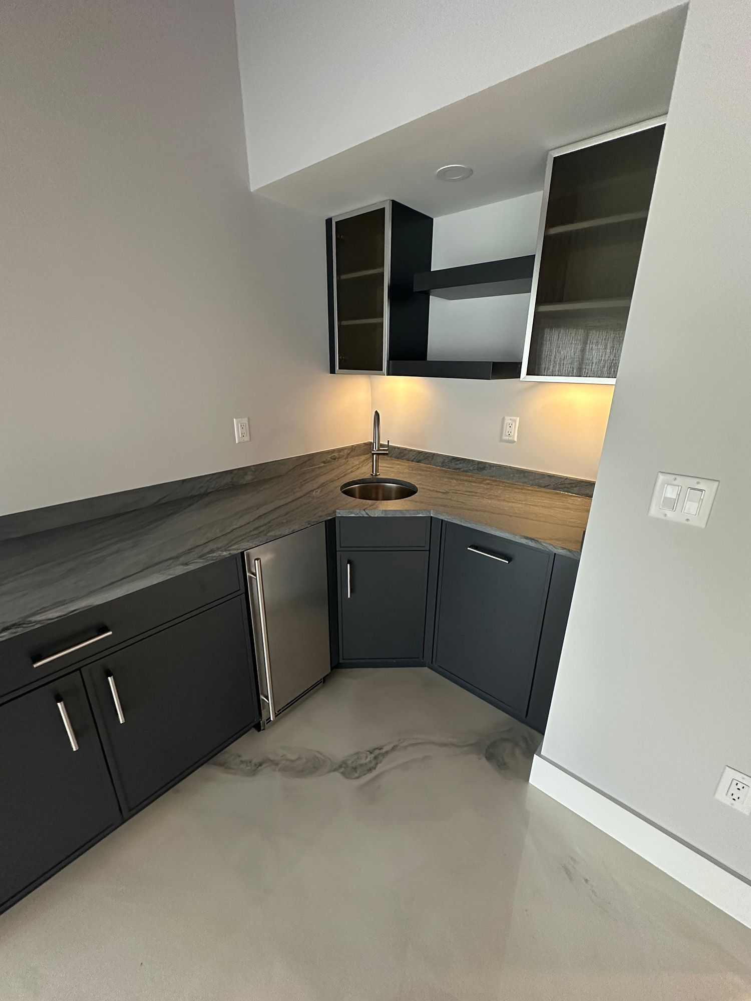 Dark gray cabinetry and countertops with a sink, mini-fridge, and overhead storage in a corner bar.