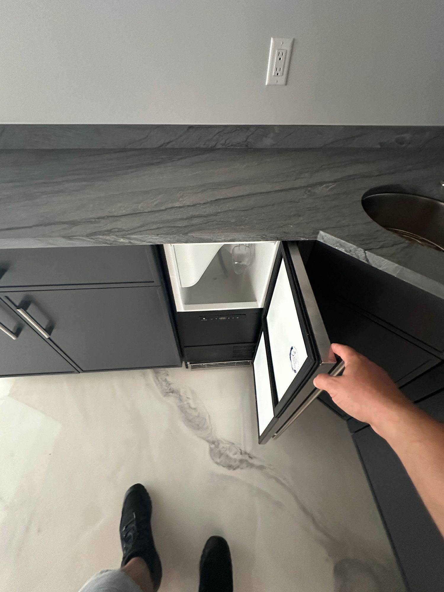 Person opening a dark cabinet door under a countertop, revealing a built-in refrigerator.