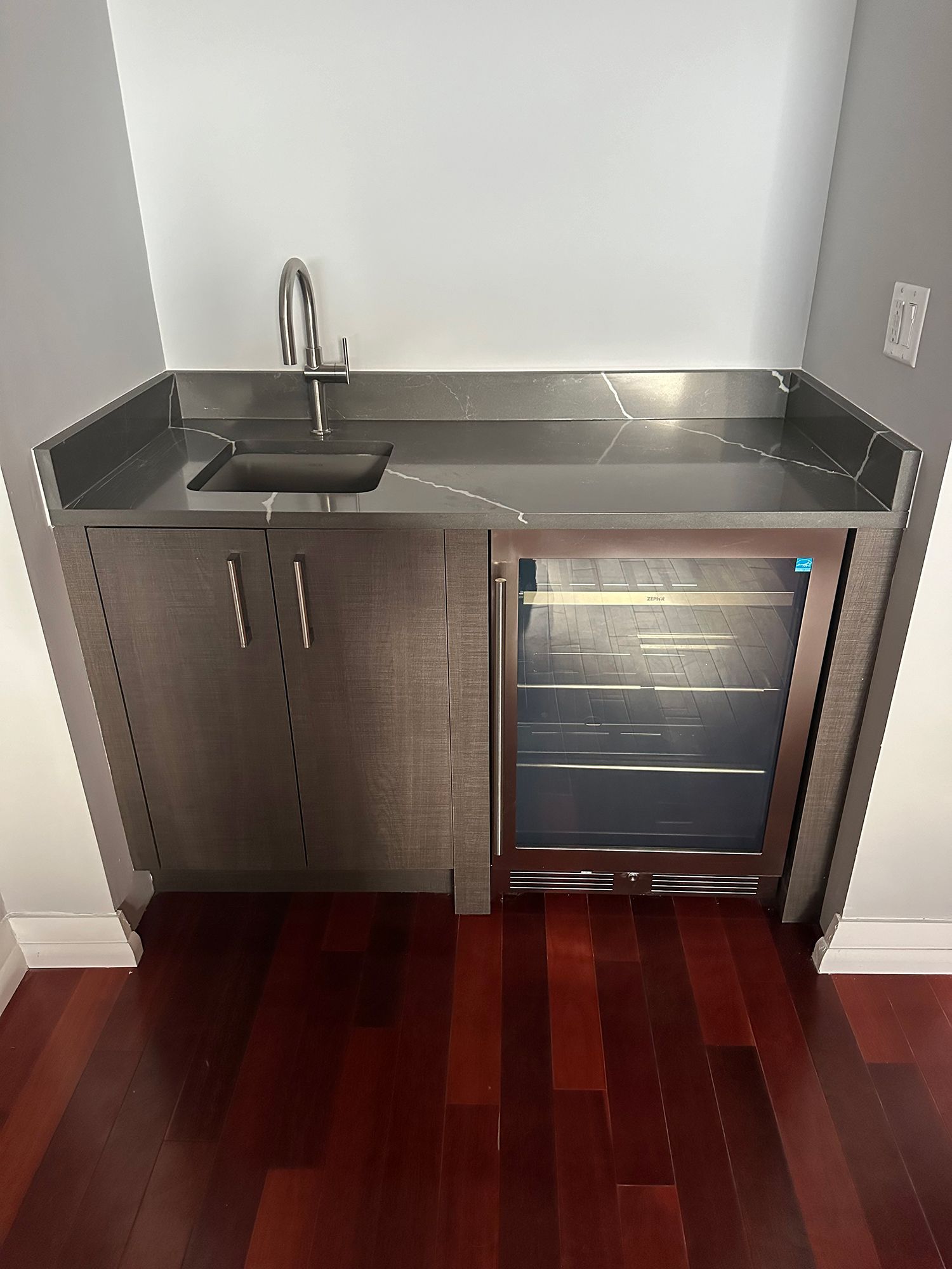Built-in bar with a sink, mini-fridge, and gray countertop against white walls and a wood floor.