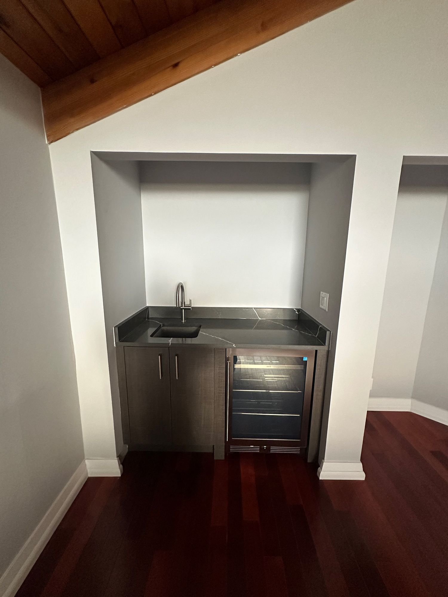 Built-in wet bar in a room with wood flooring. Stainless steel sink and mini-fridge in a recessed alcove.