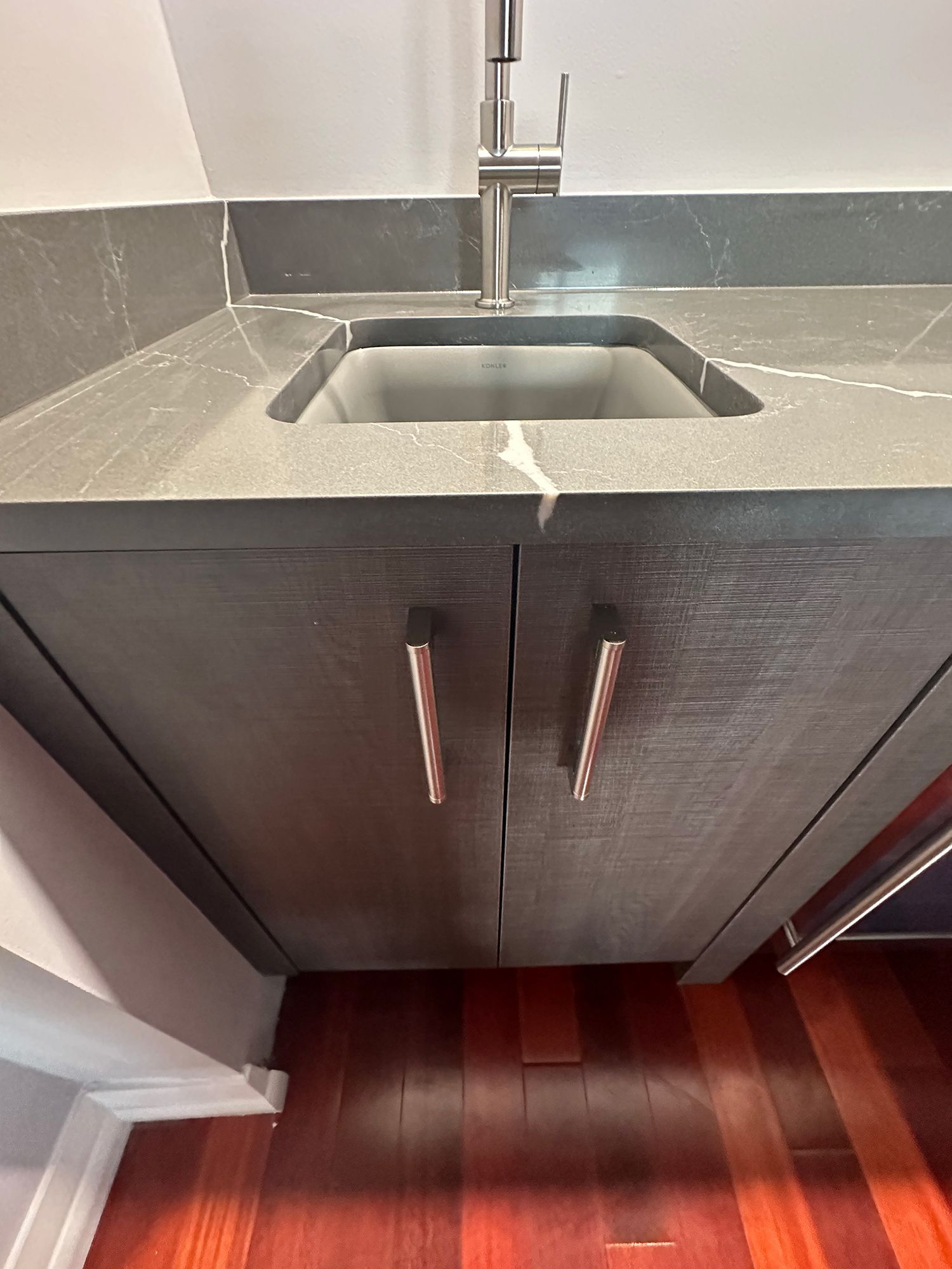 Dark cabinet with two doors, gray countertop, and stainless steel faucet and sink. Wooden floor.
