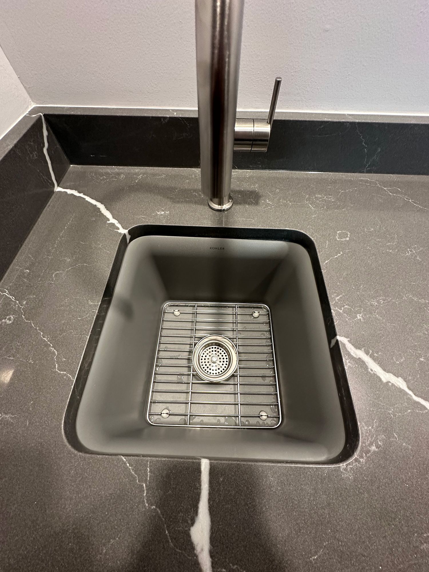 Square, dark gray kitchen sink with metal grate, stainless steel faucet. Granite countertop.