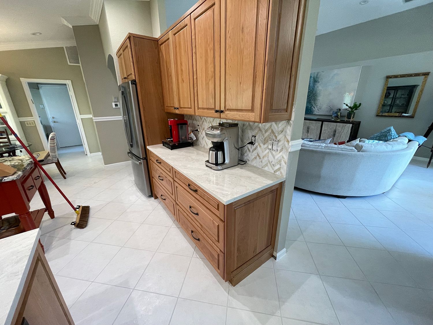 A kitchen with wood cabinets, white countertops, and white tile flooring. A refrigerator and coffee makers are visible.