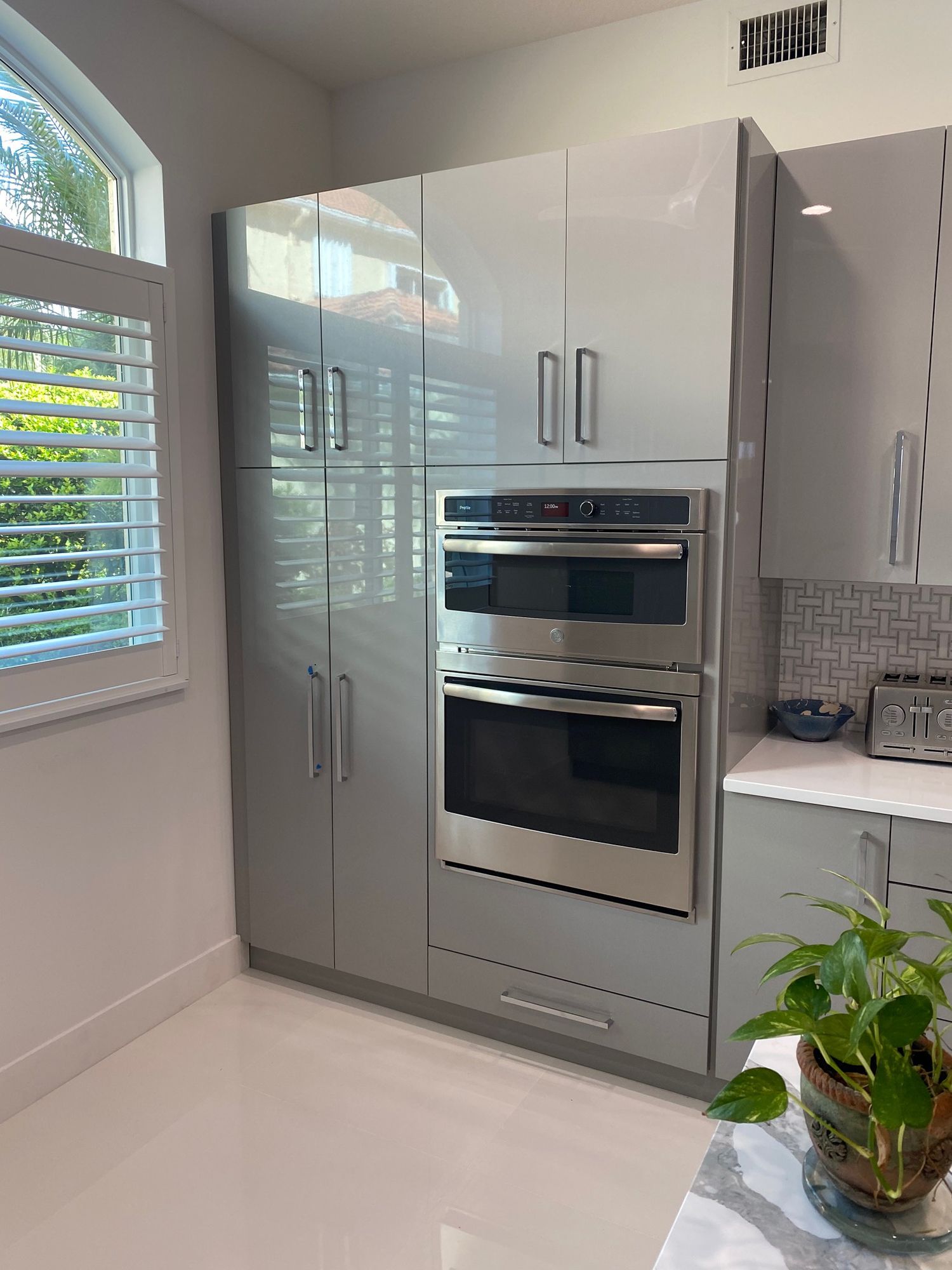 Modern gray kitchen cabinets with built-in oven and microwave, white countertop, and window with shutters.