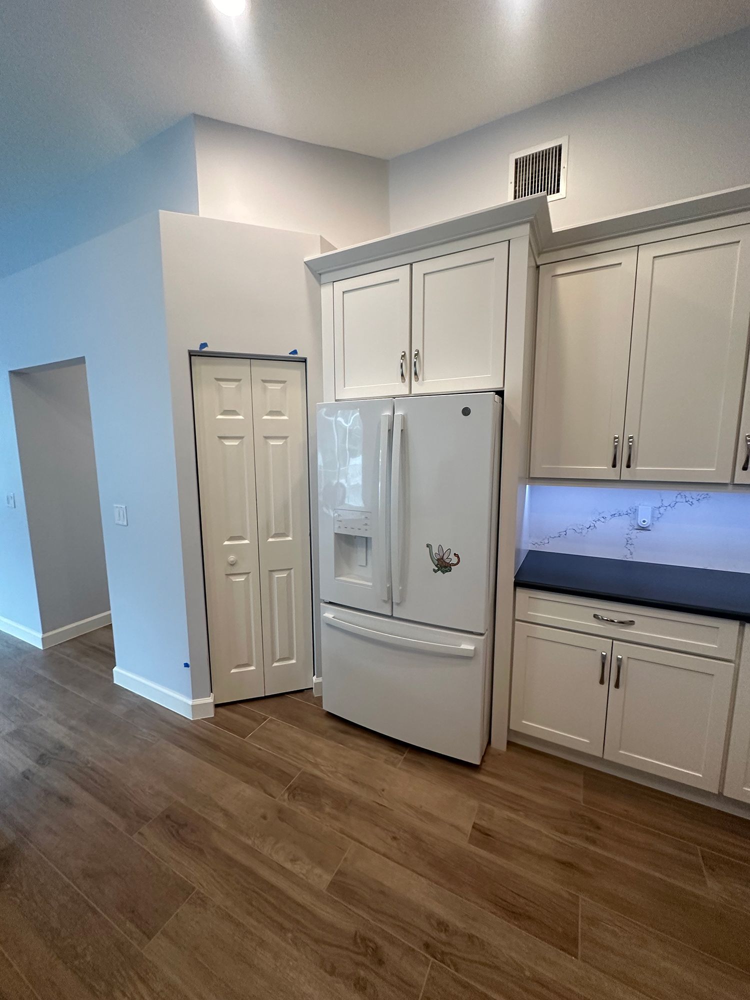 White kitchen with refrigerator, cabinets, and closed pantry door. Wooden floor.