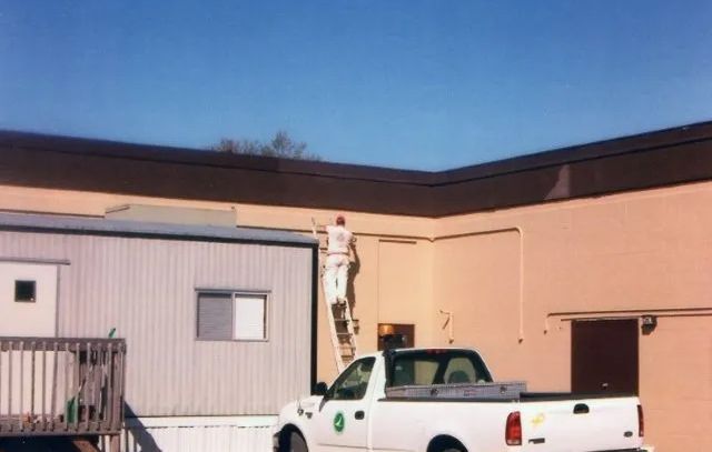 A man on a ladder paints the side of a building next to a white truck