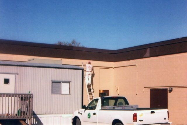 A man on a ladder paints the side of a building next to a white truck
