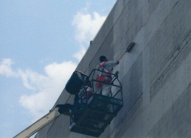 A man paints the side of a building from a crane