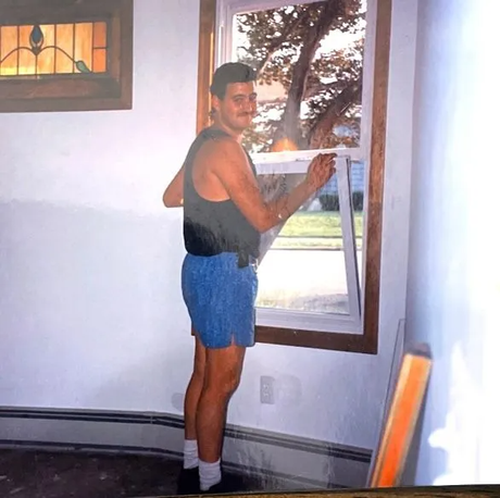 Man in blue shorts and black tank top installing a window. Interior shot with wooden trim.