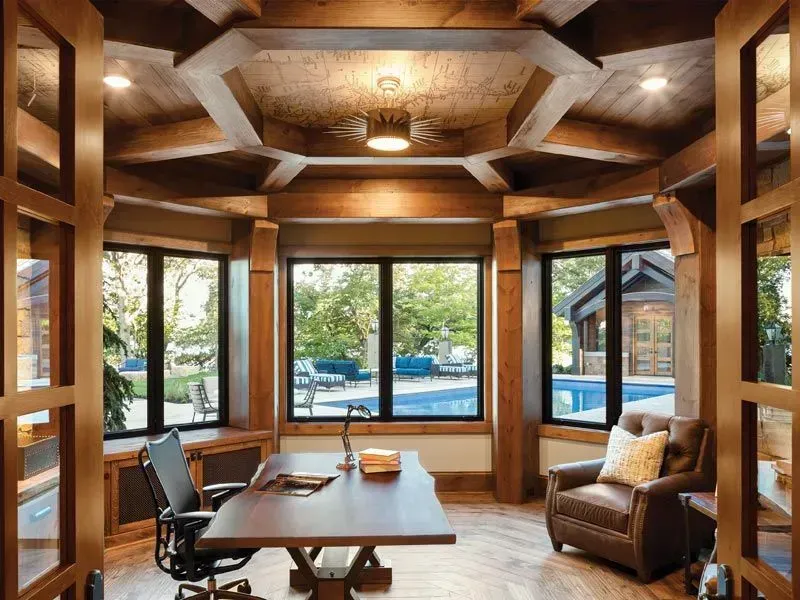 Home office with dark wood beams, large windows overlooking a pool, desk, and leather chair.